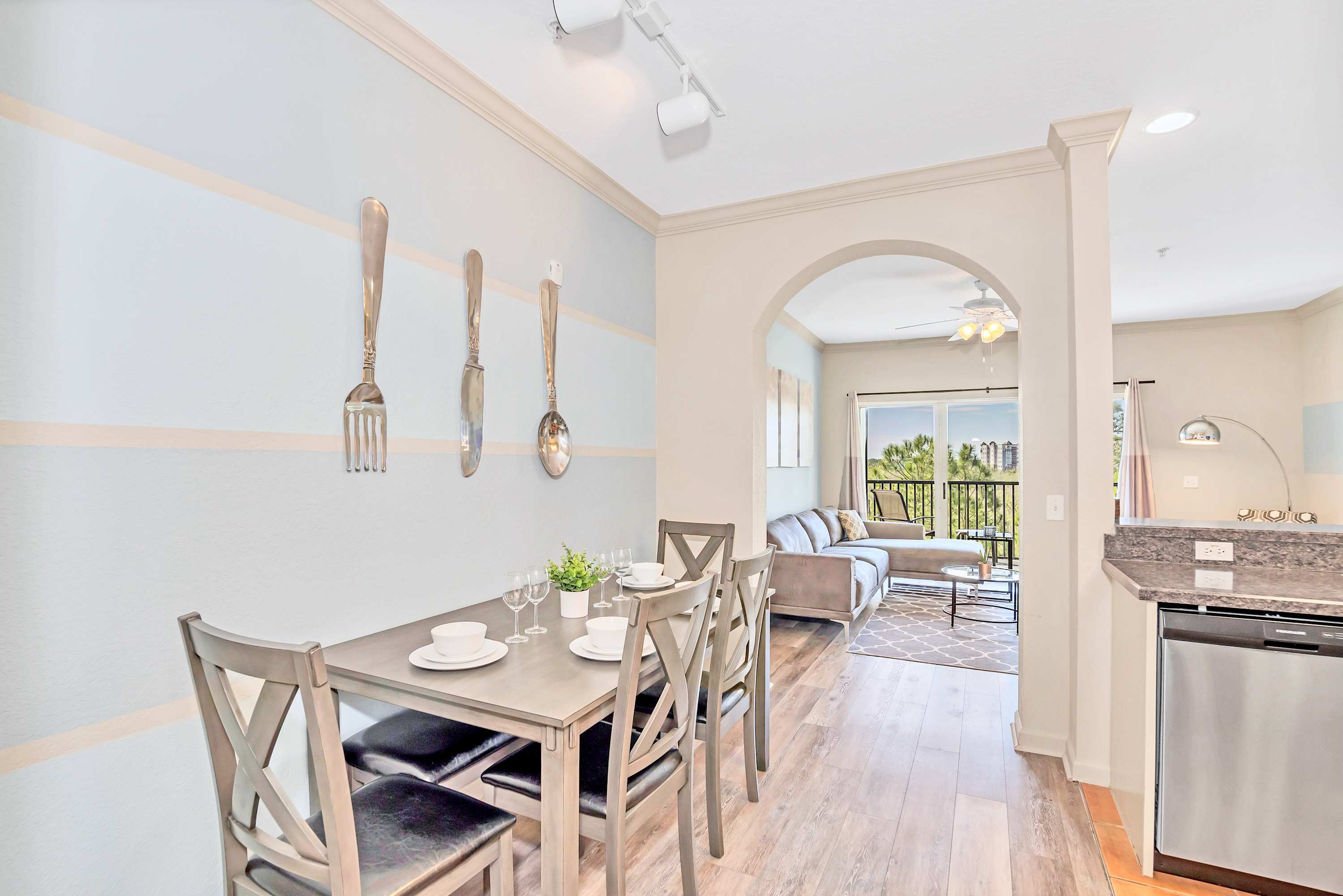 Cozy dining area featuring a rectangular wooden table set for four with matching cross-back chairs. The accent wall is painted with light blue and beige stripes, adorned with oversized metal fork, knife, and spoon wall art, with an archway leading to the living room.