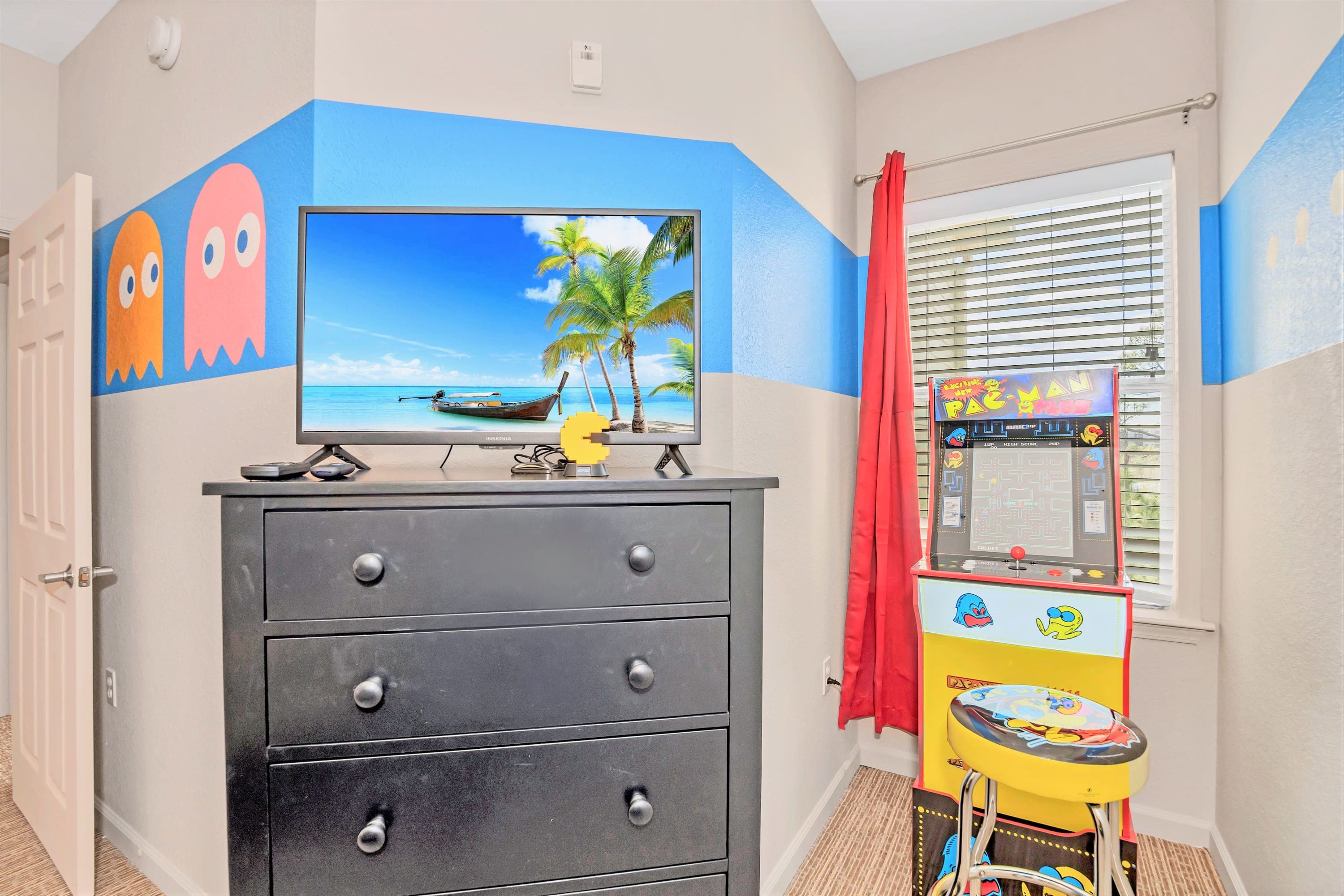 Corner of the Pac-Man themed children's bedroom featuring a black dresser with a TV, a window with blinds and red curtains, and a full-size Pac-Man arcade game machine with a matching stool.