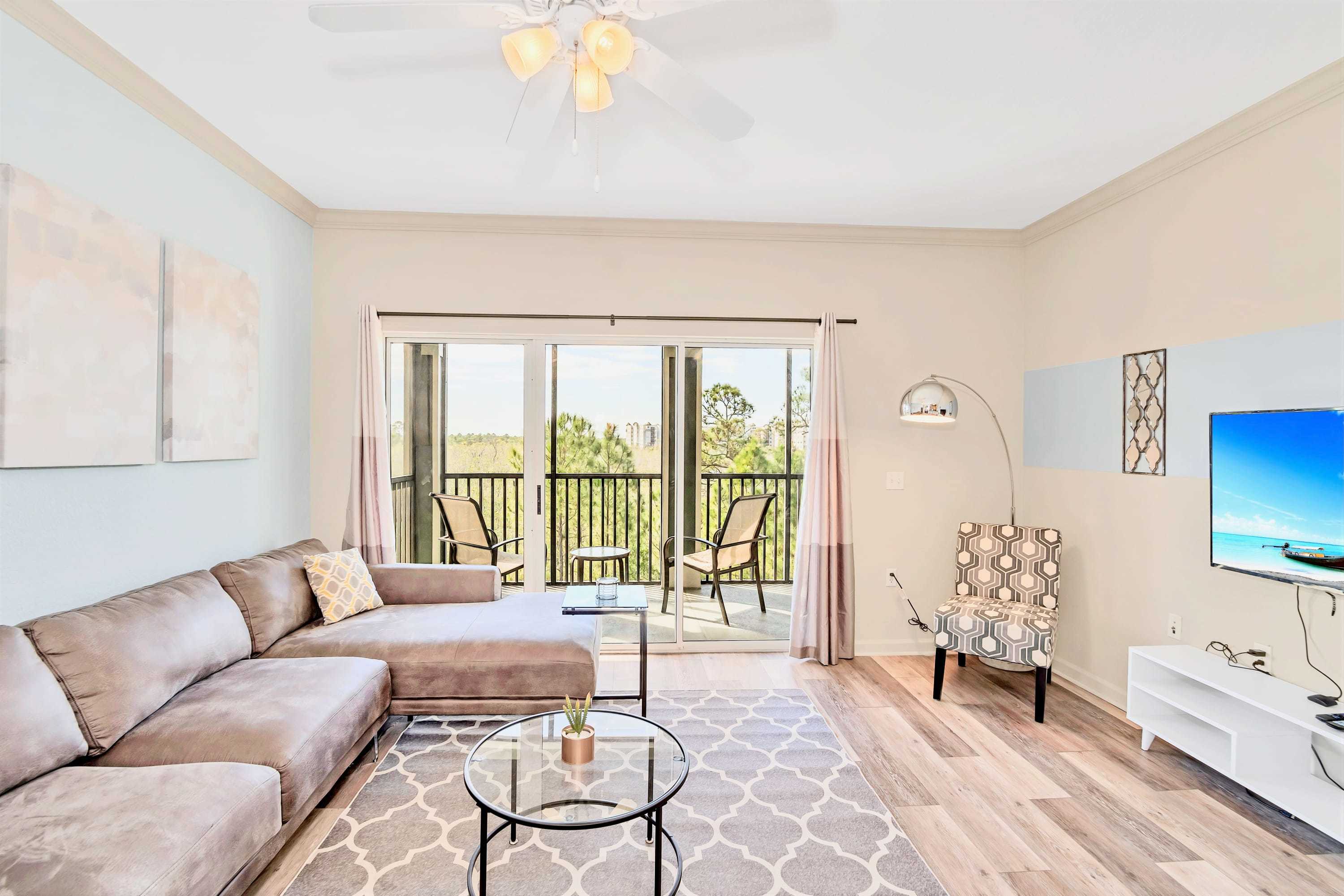 Spacious living room with a large L-shaped sectional sofa, two round glass coffee tables, and a patterned area rug on light wood-look flooring. A wall-mounted TV is visible, and sliding glass doors lead to a balcony with outdoor seating.