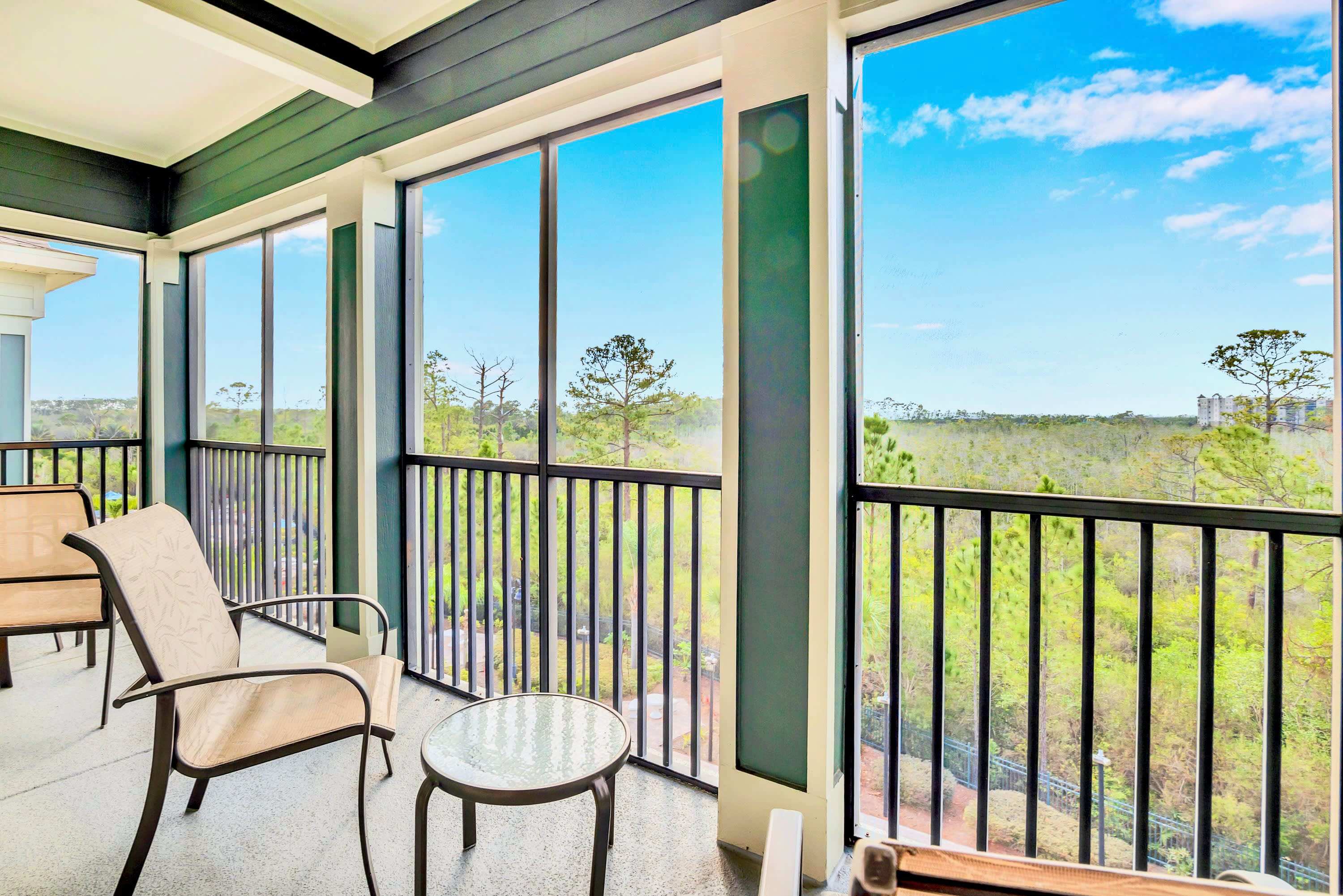 Screened-in balcony offering panoramic views of lush green trees and distant buildings, furnished with two outdoor chairs and a small round table. The space features dark green framing and a black railing.