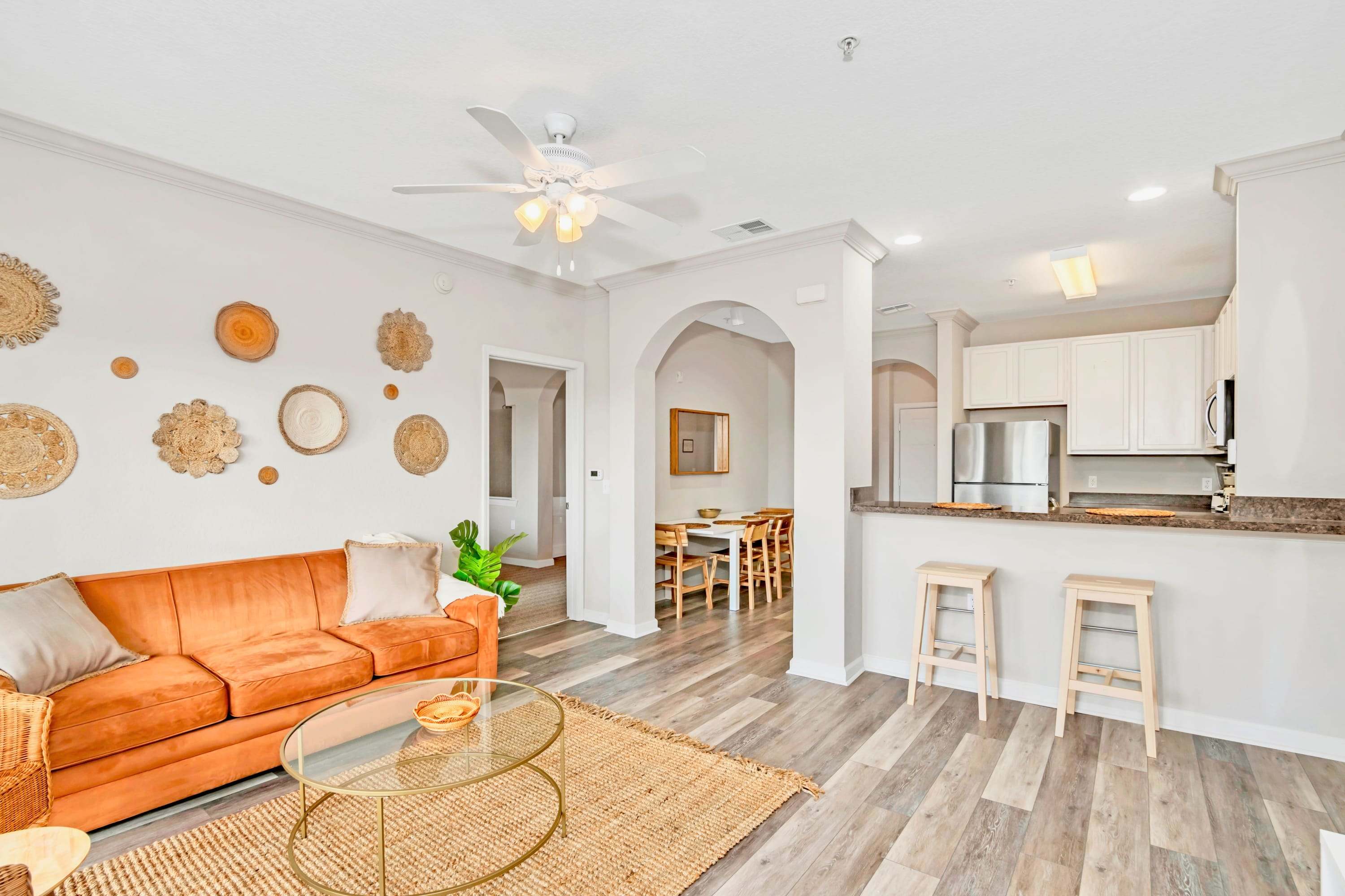 Open-concept living space featuring an orange sofa, glass coffee table on a jute rug, and a breakfast bar with two stools. An arched entryway leads to the dining area, and the kitchen is visible in the background.