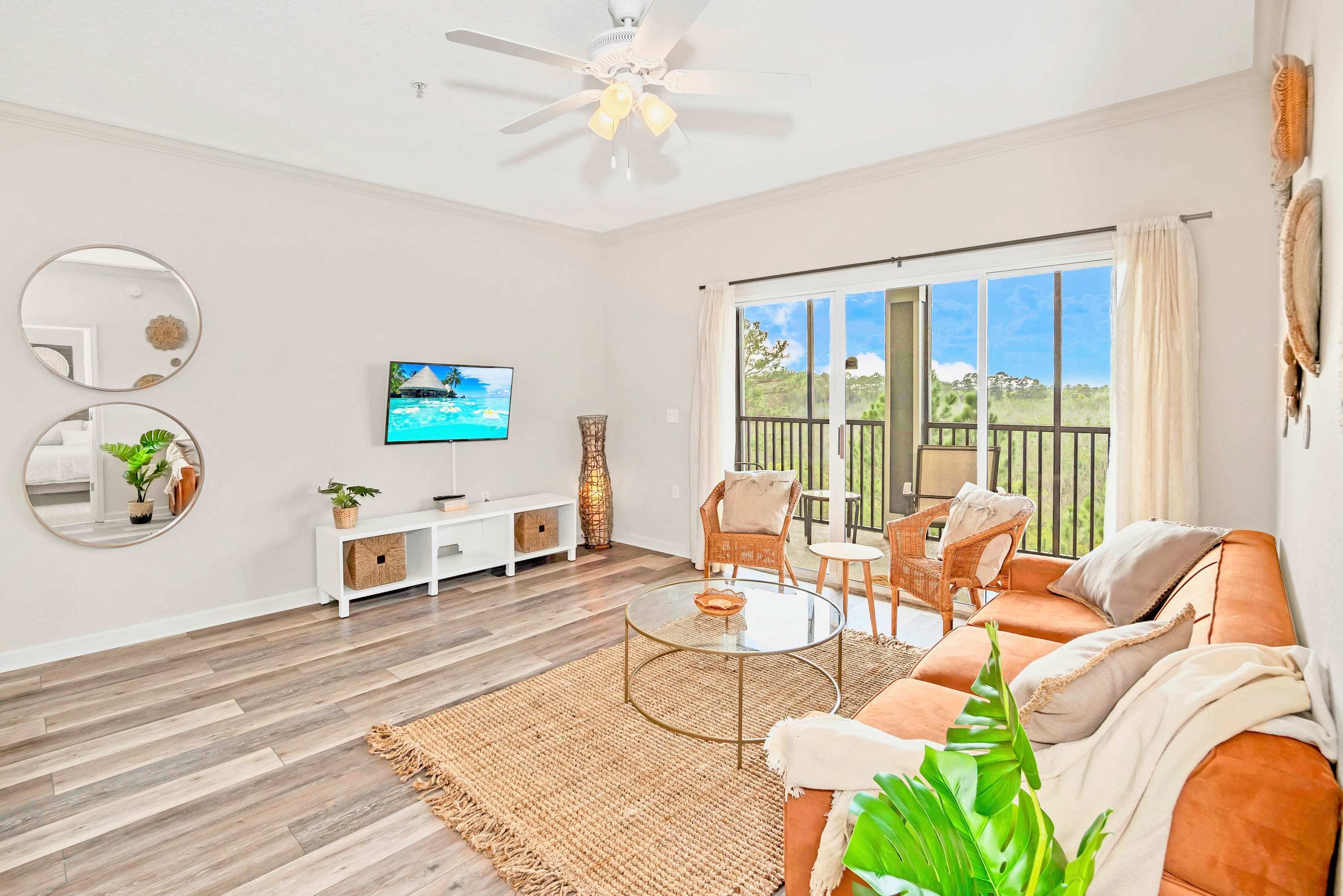 Bright and airy living room with a flat-screen TV on a white console, two round mirrors on the wall, and a view towards the balcony. The room features wood-look flooring and a ceiling fan.