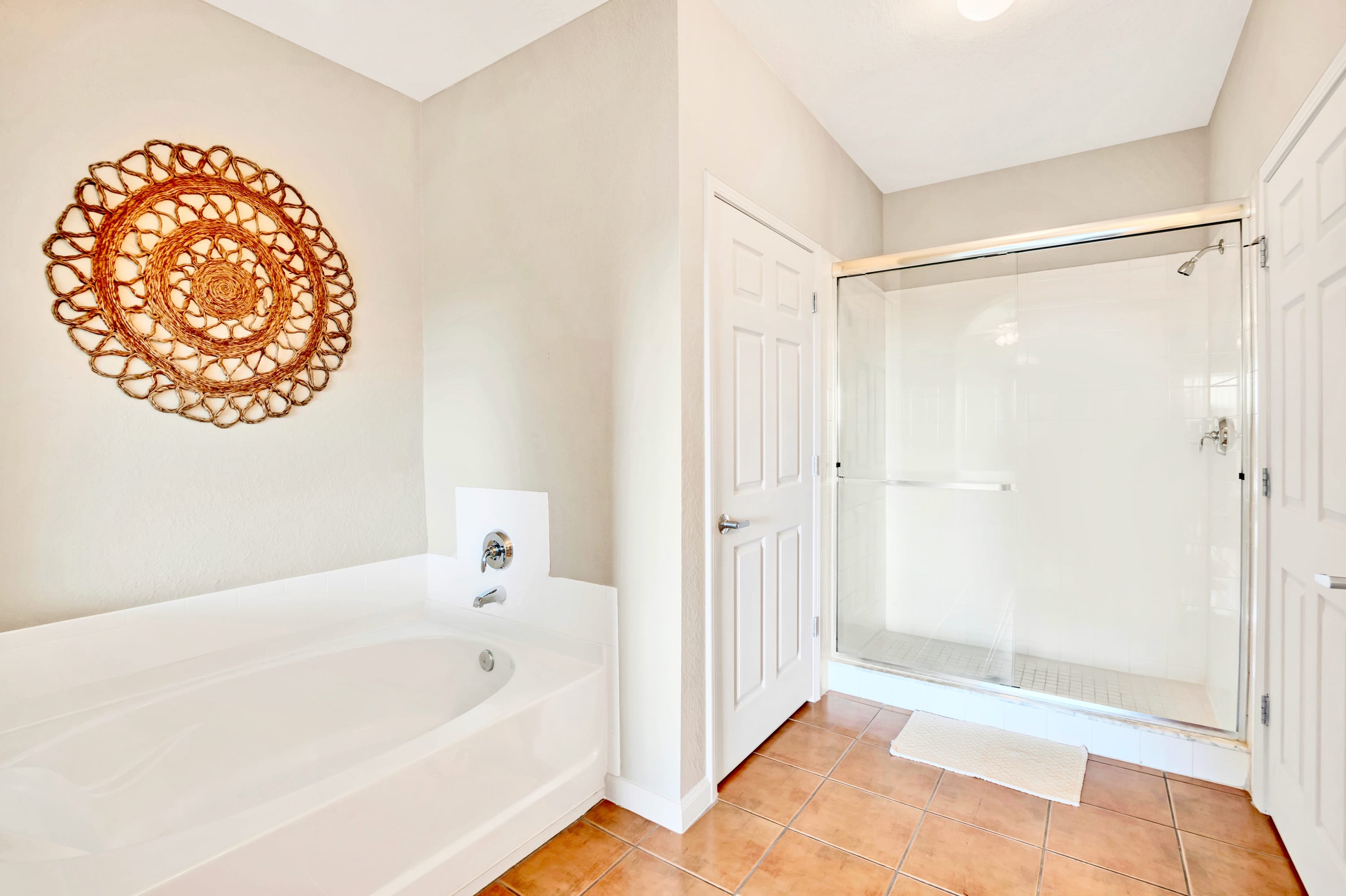 Bright bathroom area with a white soaking tub, a clear glass-enclosed shower, and light brown tiled flooring, complemented by a decorative woven wall hanging.