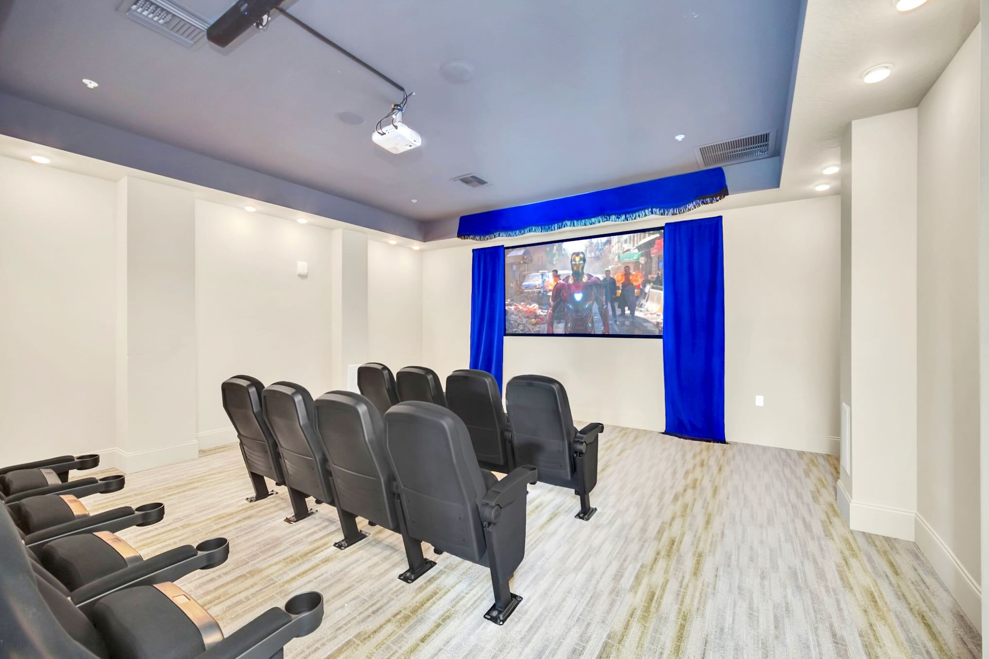 A dedicated media room features multiple rows of black cinema-style seating facing a large projection screen, framed by blue curtains, with a projector mounted on the ceiling.