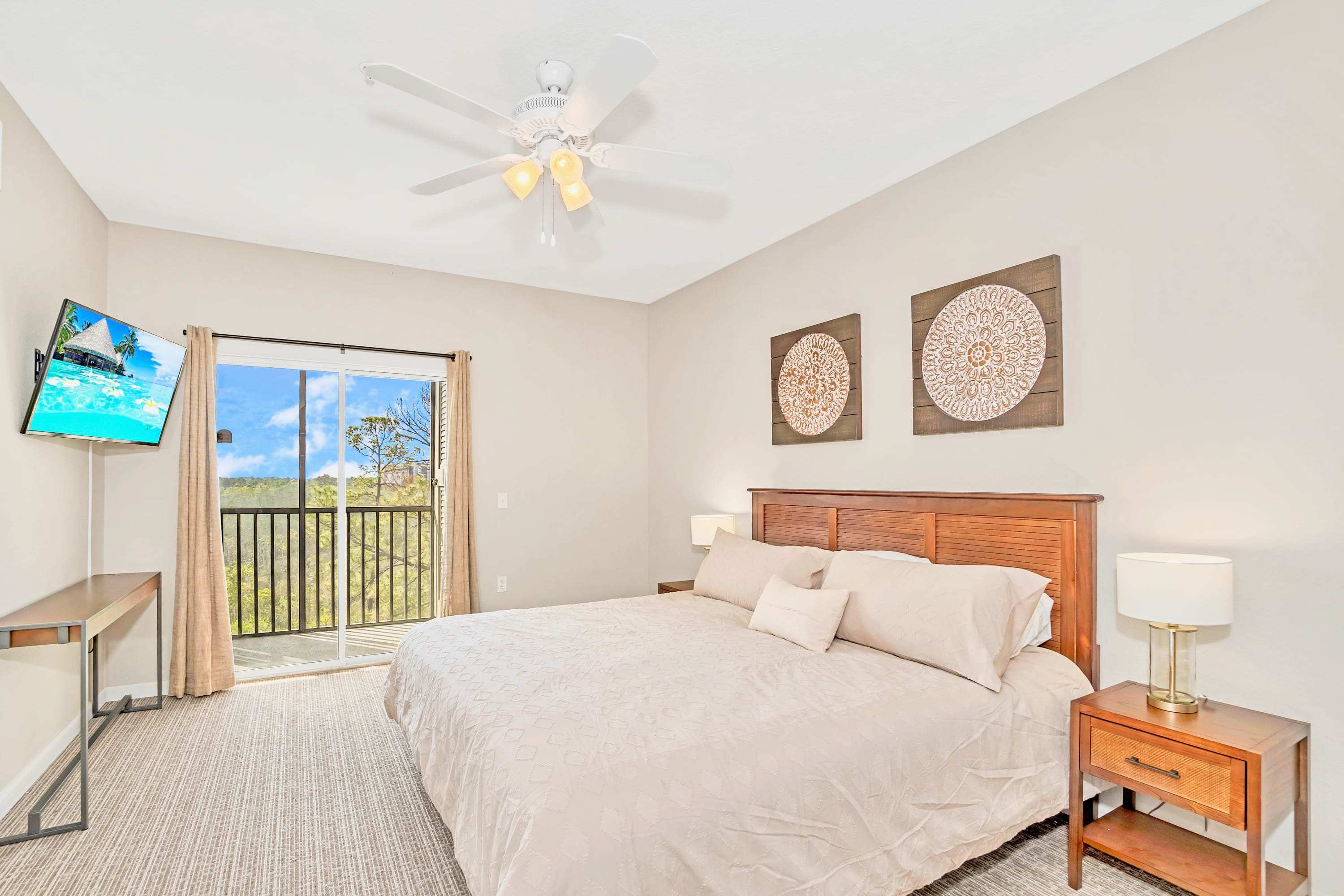 Spacious bedroom with a wooden king-sized bed, two nightstands with lamps, a wall-mounted TV, and a private balcony overlooking lush greenery, featuring carpeted flooring and a ceiling fan.