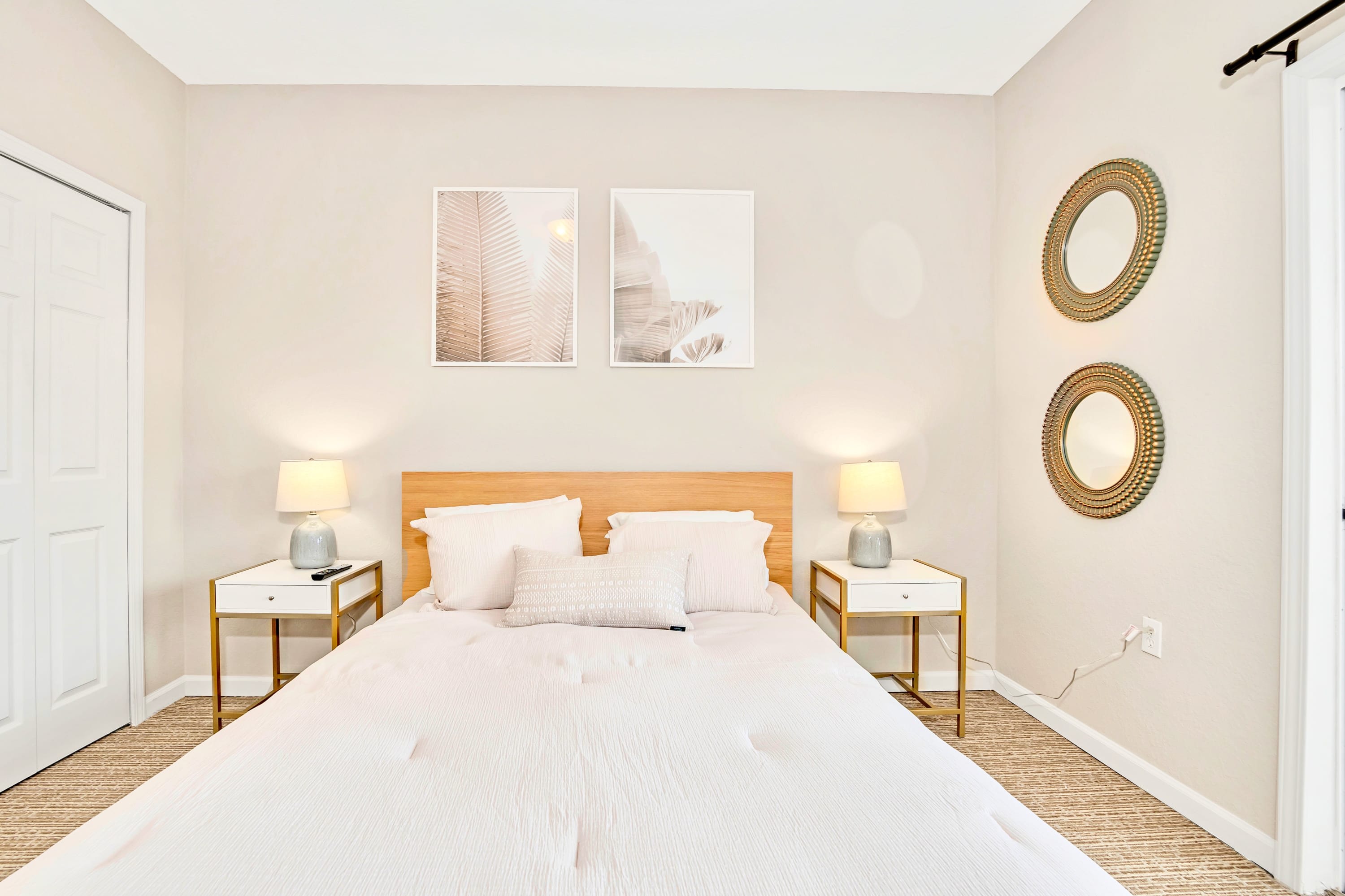 Close-up view of the queen bed in bedroom-2, featuring a light wood headboard, two nightstands with lamps, and two framed botanical prints above the bed, with closet doors visible.