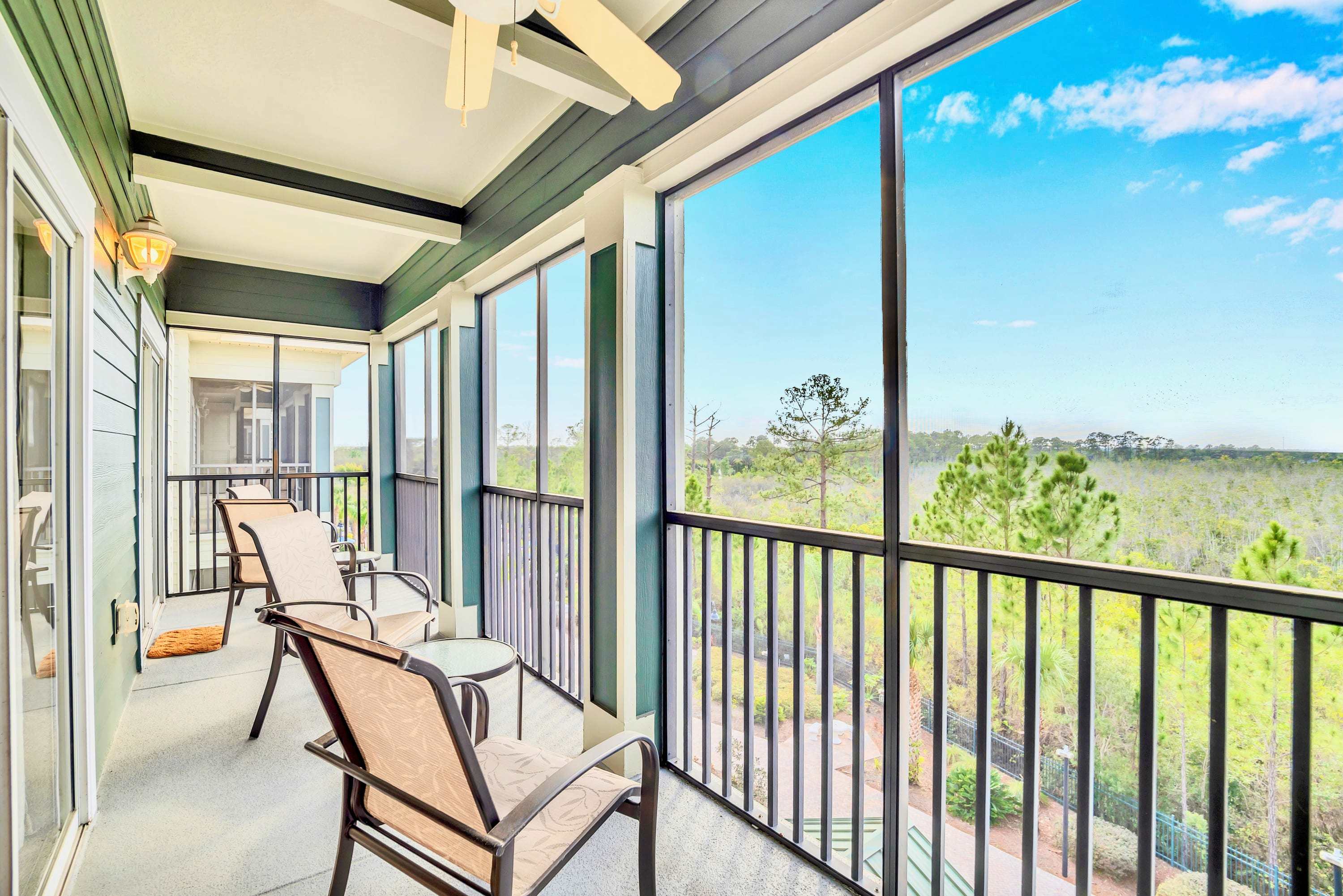 Screened-in balcony with outdoor seating, including two chairs and a small table, offering expansive views of a natural, tree-filled landscape under a bright blue sky. The ceiling has a fan and recessed lighting.
