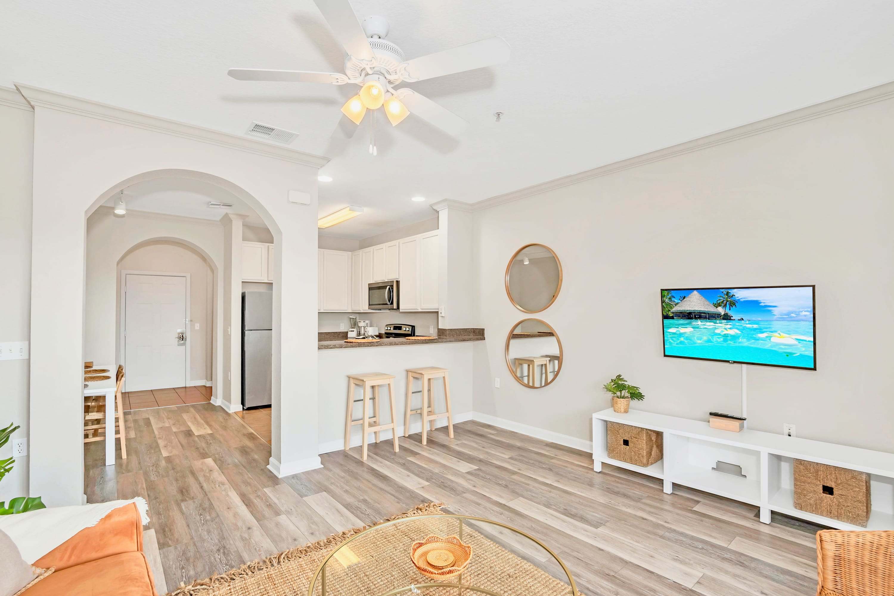 Bright living room with wood-look flooring, a ceiling fan, and a flat-screen TV, opening to a kitchen bar area with two stools and an arched hallway leading to other rooms.