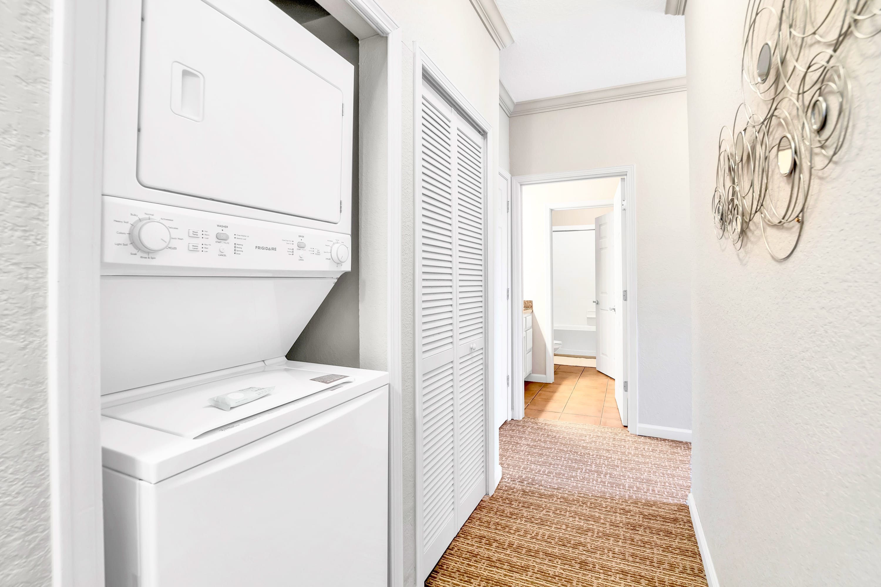 A narrow hallway features a stacked white washer and dryer unit on the left, with a closed louvered door closet on the right, leading to a bathroom visible in the background.