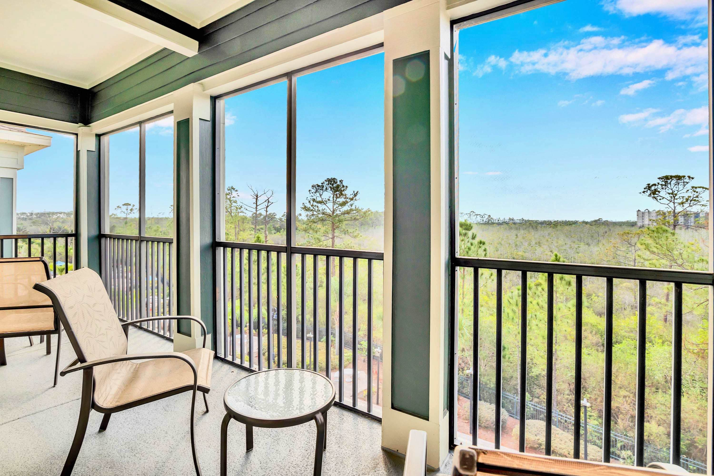 Screened-in balcony with a small round table and two chairs, offering a panoramic view of a vast, natural landscape with dense trees under a clear blue sky. The balcony has dark railings and a ceiling fan.