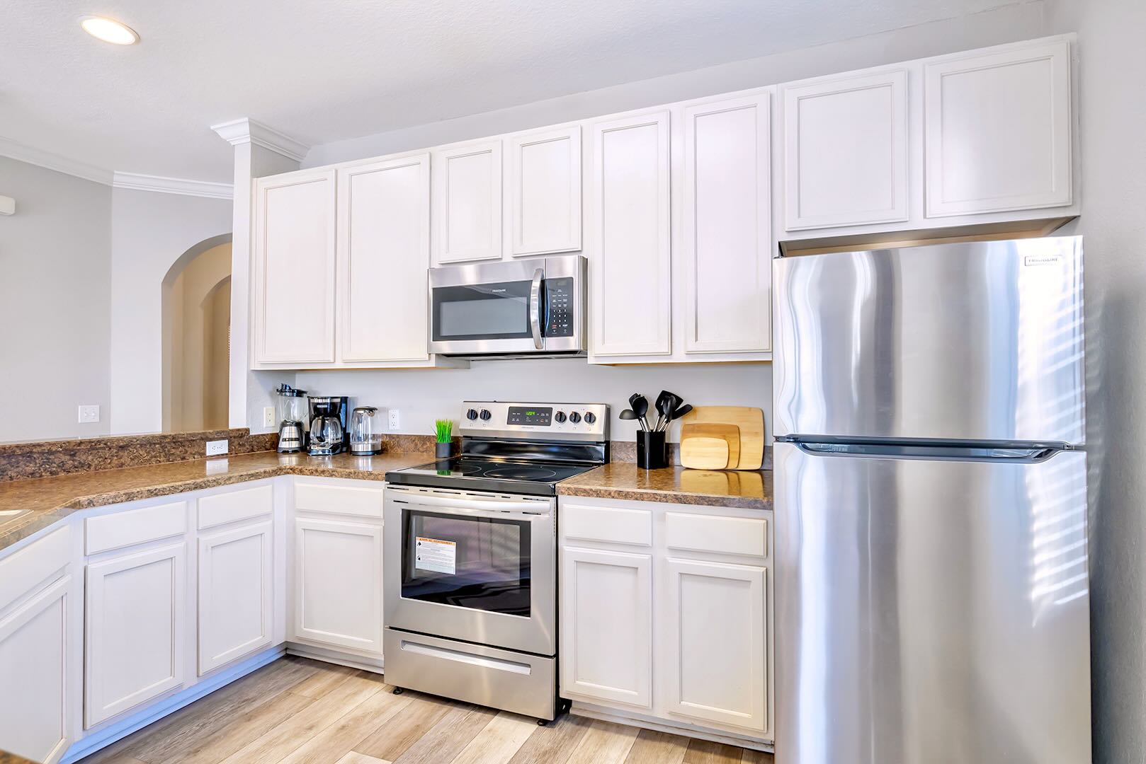 Close-up view of the kitchen showing white shaker cabinets, stainless steel microwave, electric range, and a large stainless steel refrigerator. The brown speckled countertop holds a coffee maker and various kitchen utensils.