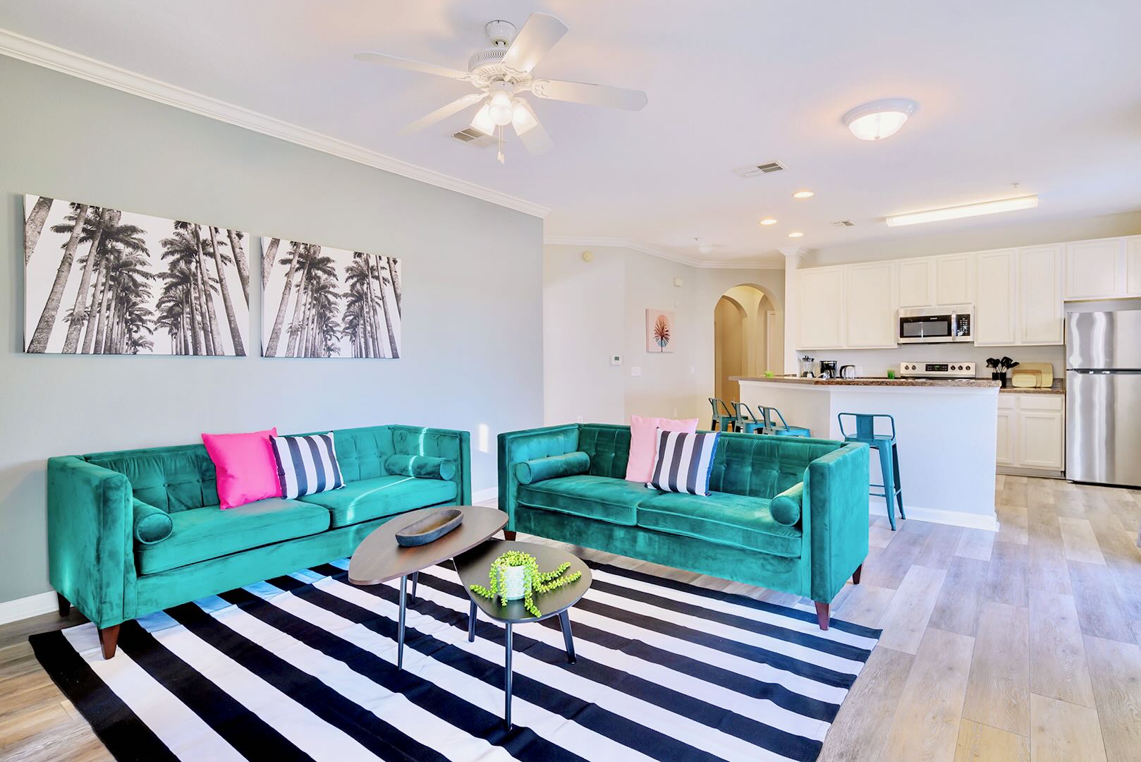 Wide shot of an open-concept living room featuring two teal velvet sofas with pink and striped pillows, a black and white striped rug, and modern coffee tables. The kitchen with white cabinets is visible in the background.