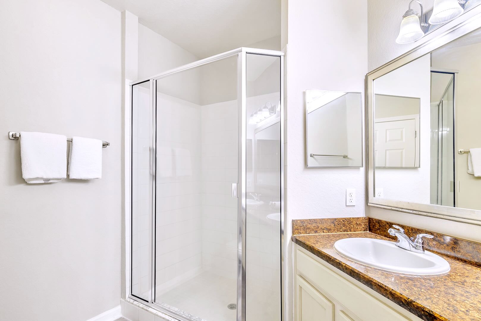 En-suite bathroom vanity area with a single sink set in a brown speckled countertop, white cabinets, and a large mirror. A glass-enclosed shower with white tile is visible next to the vanity.