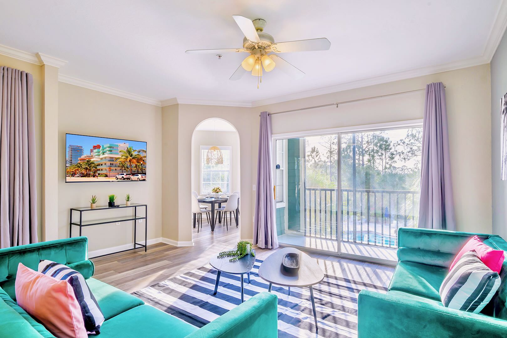 View of the living room from a different angle, showing two teal sofas, a flat-screen TV mounted on the wall, and a sliding glass door leading to a balcony. An arched doorway leads to the dining area.