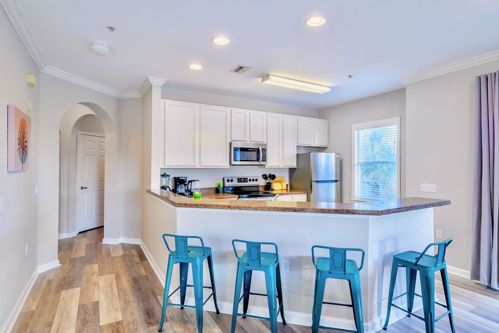 Modern kitchen with white shaker cabinets, stainless steel appliances, and a brown speckled countertop. A breakfast bar with four teal bar stools extends into the living area, featuring light wood-look flooring.