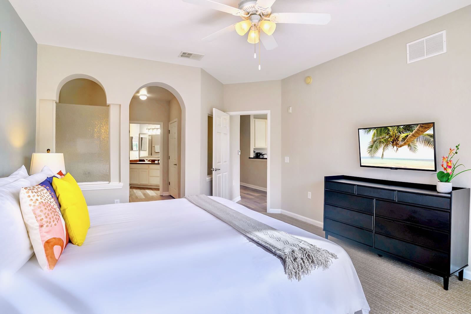 View of the master bedroom showing the bed with colorful pillows, a black dresser with a TV, and an arched doorway leading to the en-suite bathroom. The room has light carpet and a ceiling fan.