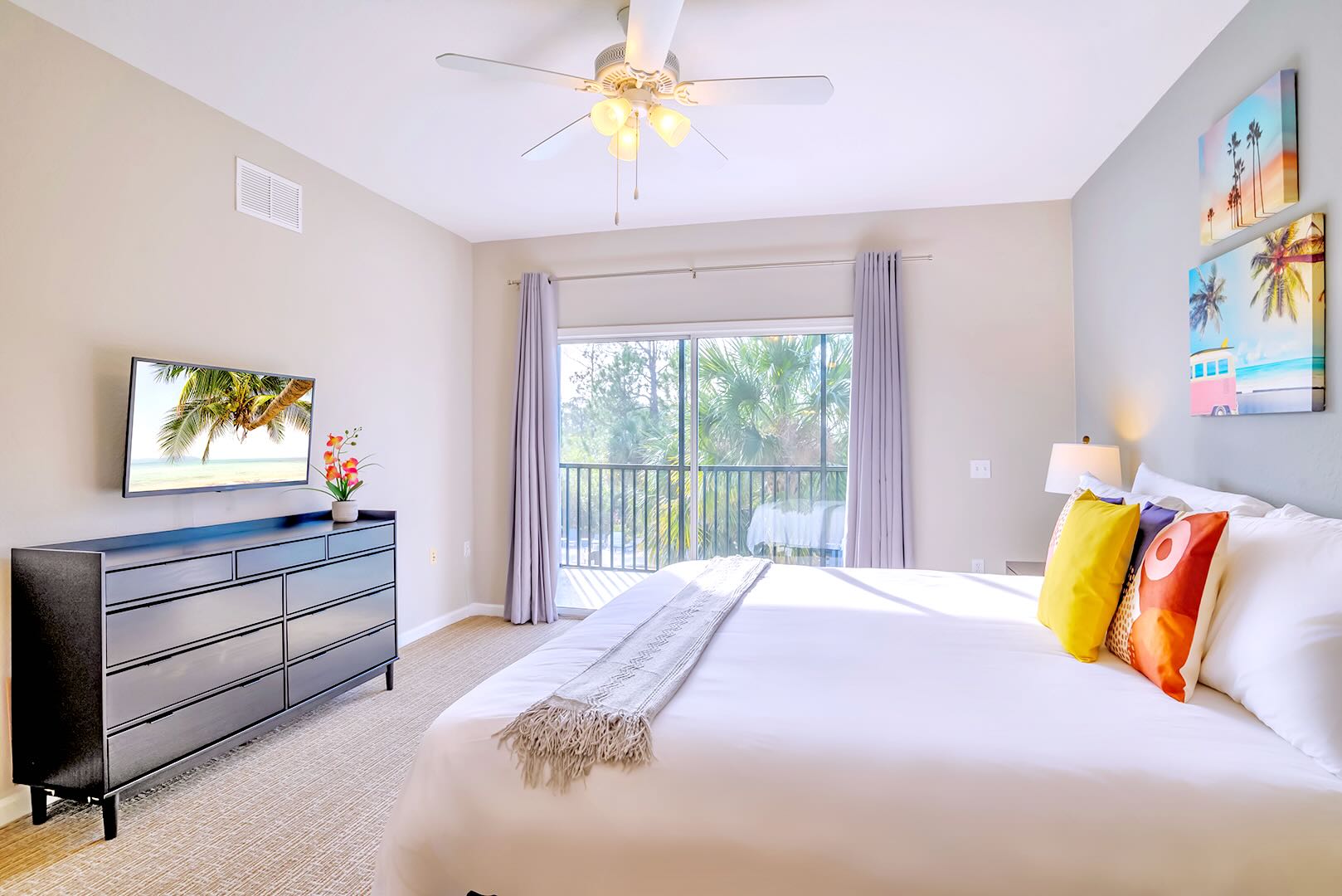 Bright bedroom with a large white bed, a black dresser with a flat-screen TV, and a sliding glass door leading to a balcony. The room features light-colored carpet and a ceiling fan.