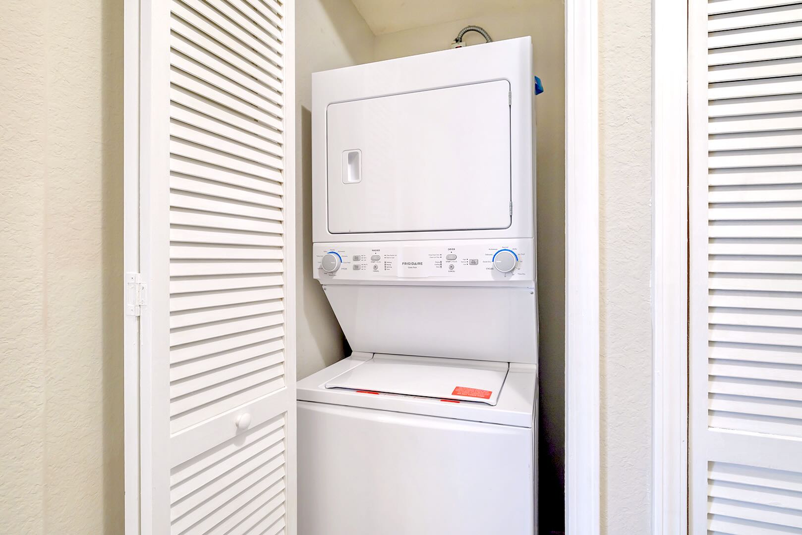 A white stacked washer and dryer unit is neatly tucked into a closet with white louvered bifold doors, featuring blue control knobs and a clean, functional design.