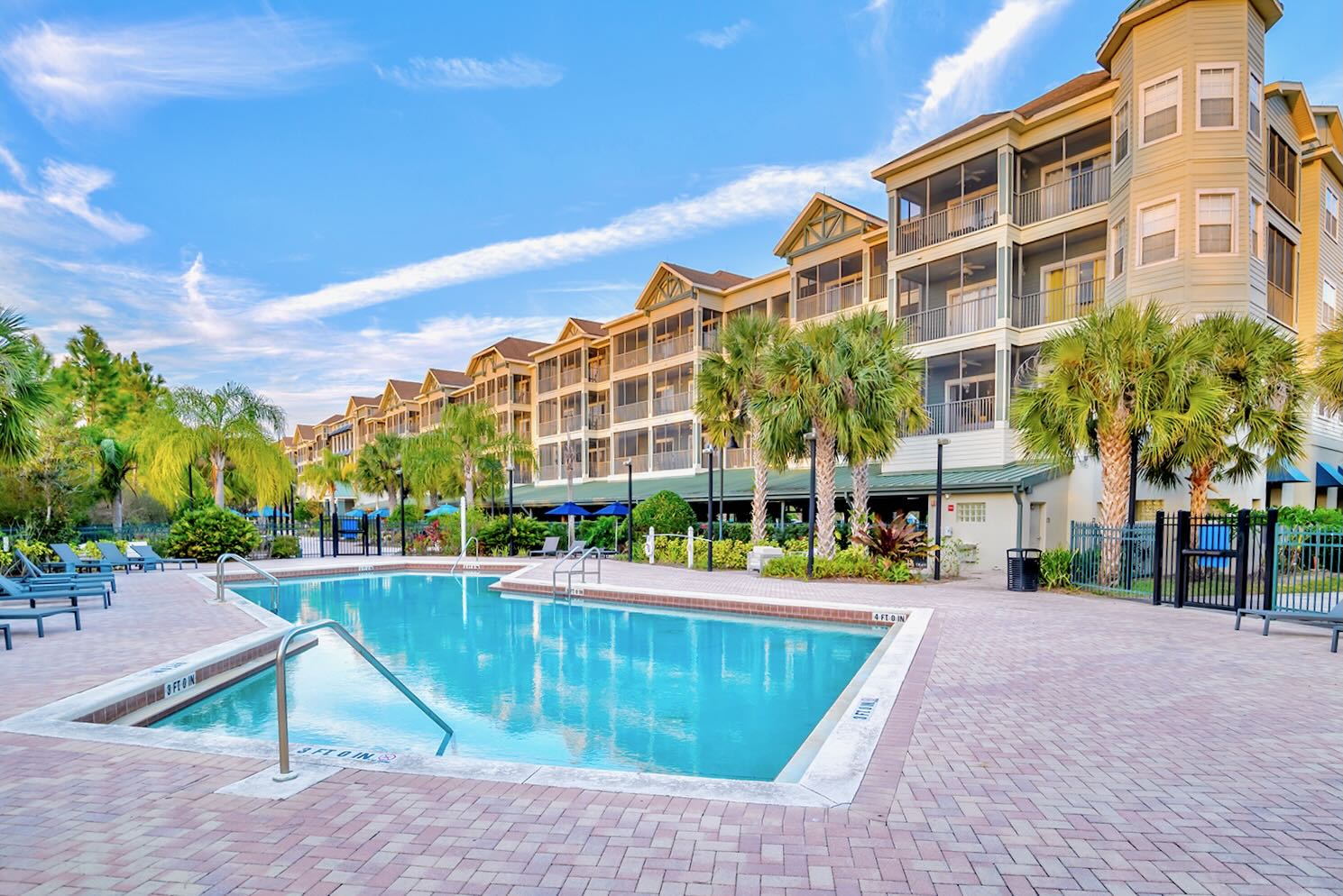 A large, rectangular outdoor swimming pool with clear blue water is surrounded by a brick paver deck, with a multi-story apartment building and palm trees in the background under a bright sky.