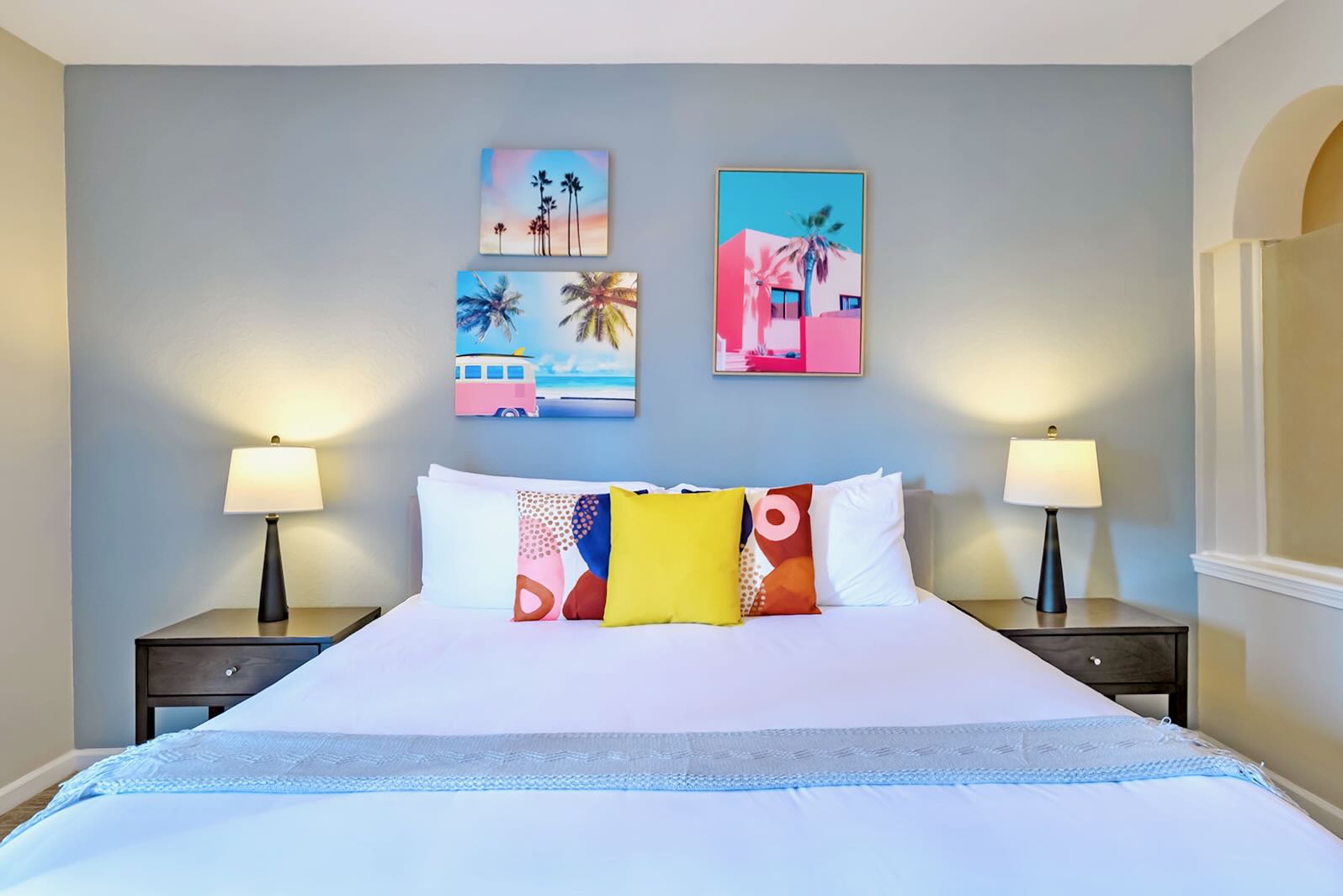 Master bedroom featuring a large bed with white bedding and colorful accent pillows, flanked by two dark wood nightstands with lamps. The light blue accent wall is decorated with three vibrant tropical-themed art pieces.
