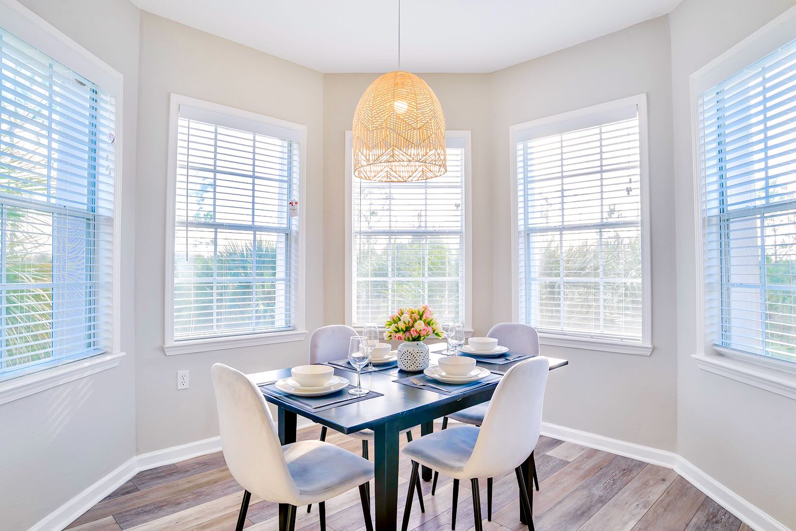 Bright dining area with a dark rectangular table and four light gray upholstered chairs, illuminated by a large woven pendant light. Multiple windows with blinds offer abundant natural light.