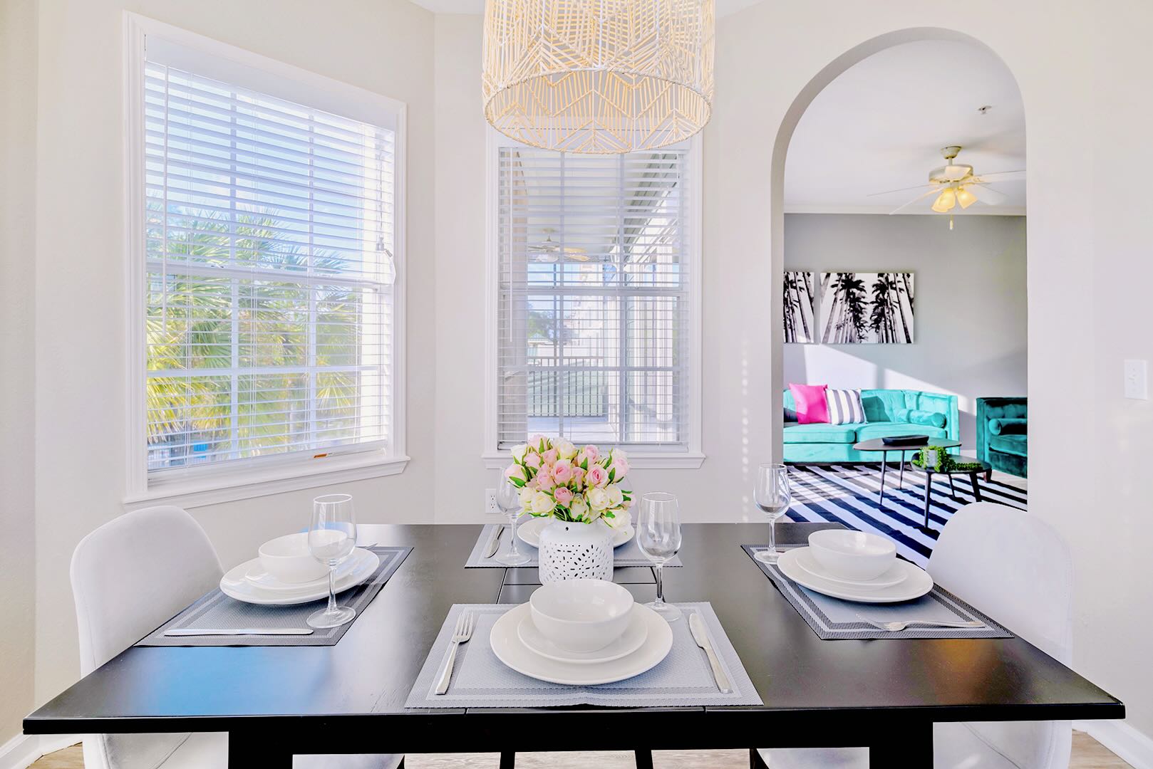Close-up view of the dining area with a dark table set for four, featuring a floral centerpiece and a large woven pendant light. An arched doorway provides a glimpse into the living room with teal sofas.
