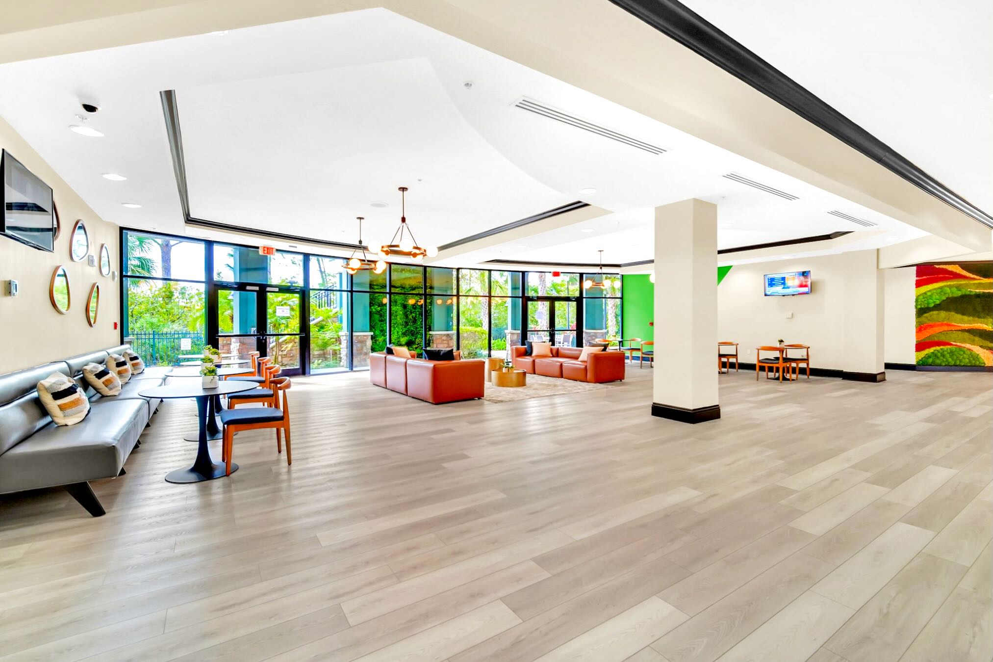 A bright and expansive communal lobby or lounge area featuring light wood laminate flooring, high ceilings with modern lighting, and floor-to-ceiling windows offering views of lush outdoor greenery. The space is furnished with various seating arrangements, including grey and orange sofas, small tables, and a wall-mounted TV, complemented by vibrant abstract artwork.
