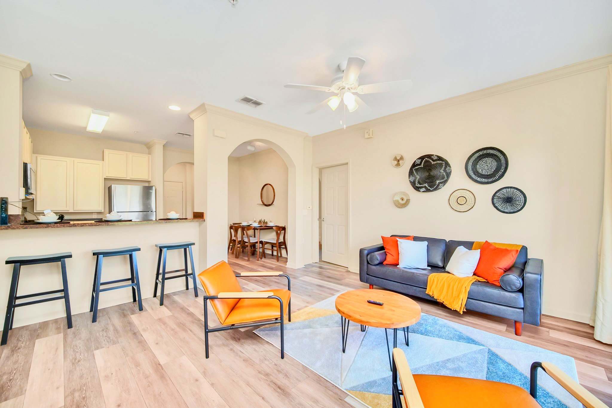 Spacious living room with light wood-look flooring, a dark leather sofa with orange pillows, two orange armchairs, and a round wooden coffee table on a geometric rug. The room connects to the kitchen and dining areas.