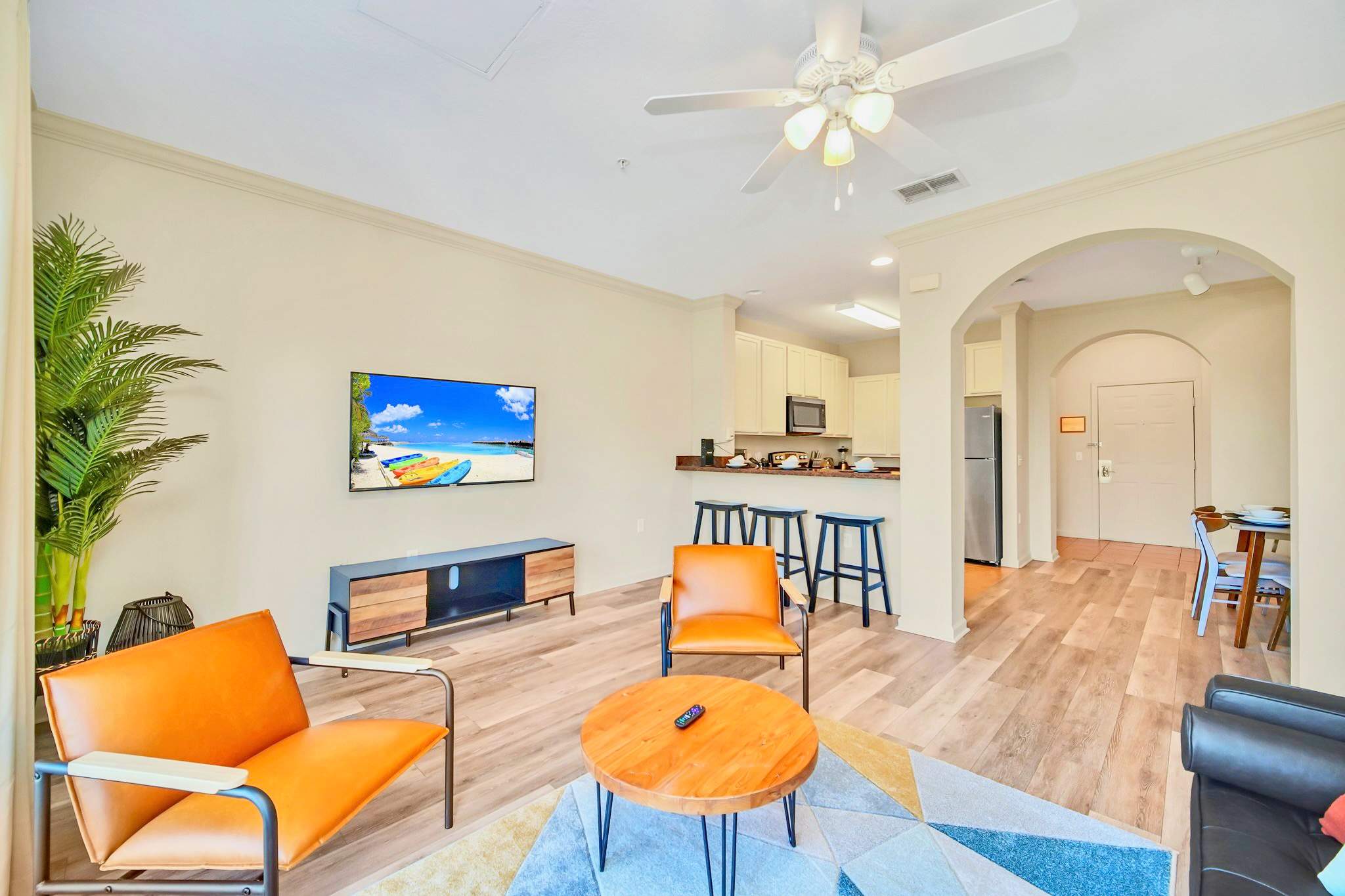 Open-concept living room with light wood-look flooring, a dark leather sofa, two orange armchairs, and a round wooden coffee table on a geometric rug. The space opens to a kitchen bar area with bar stools.
