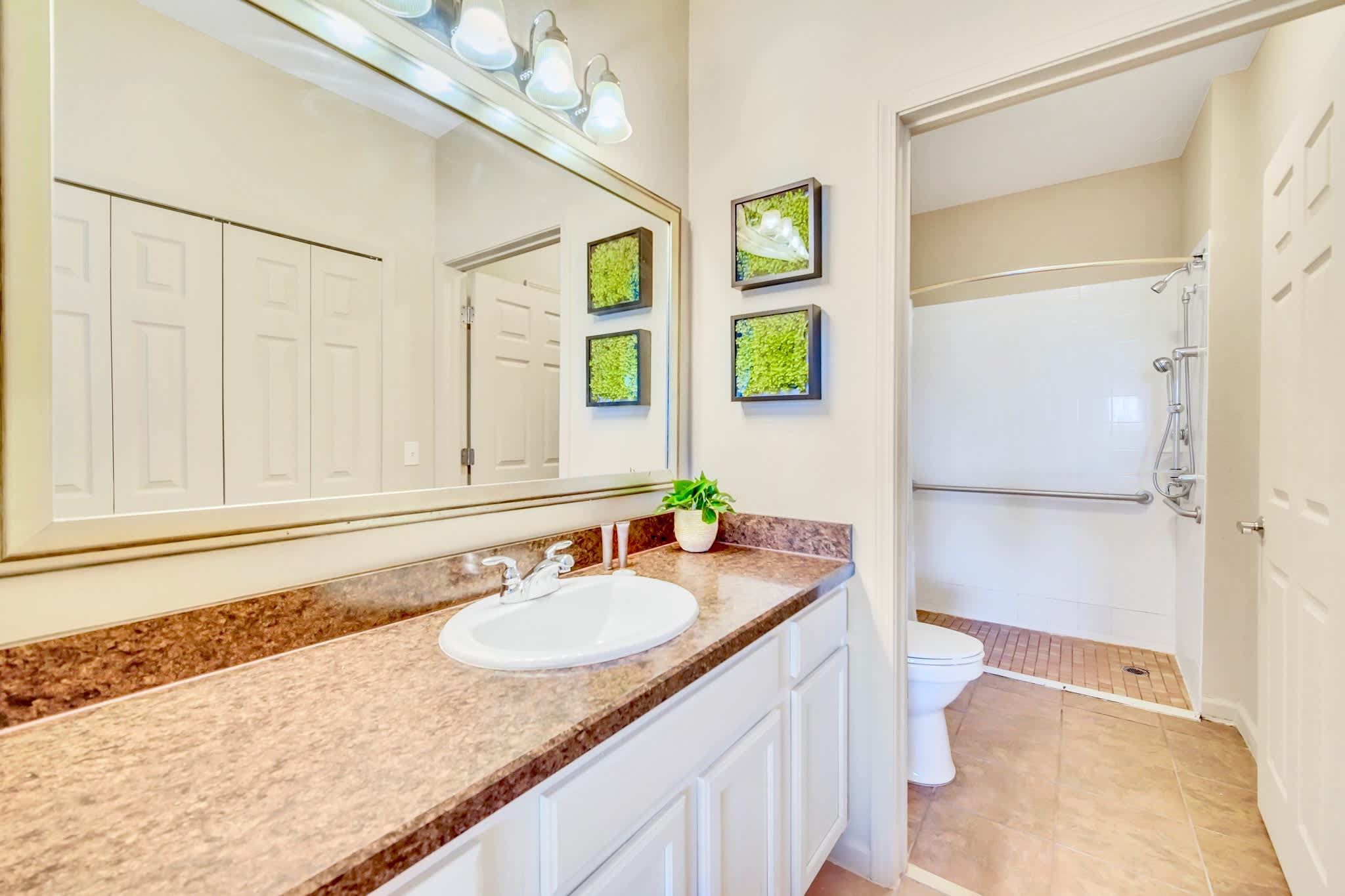 A clean bathroom showcases a single vanity with a large mirror, a light brown speckled countertop, and white shaker-style cabinets. A walk-in shower with a tiled floor and grab bars is visible to the right.