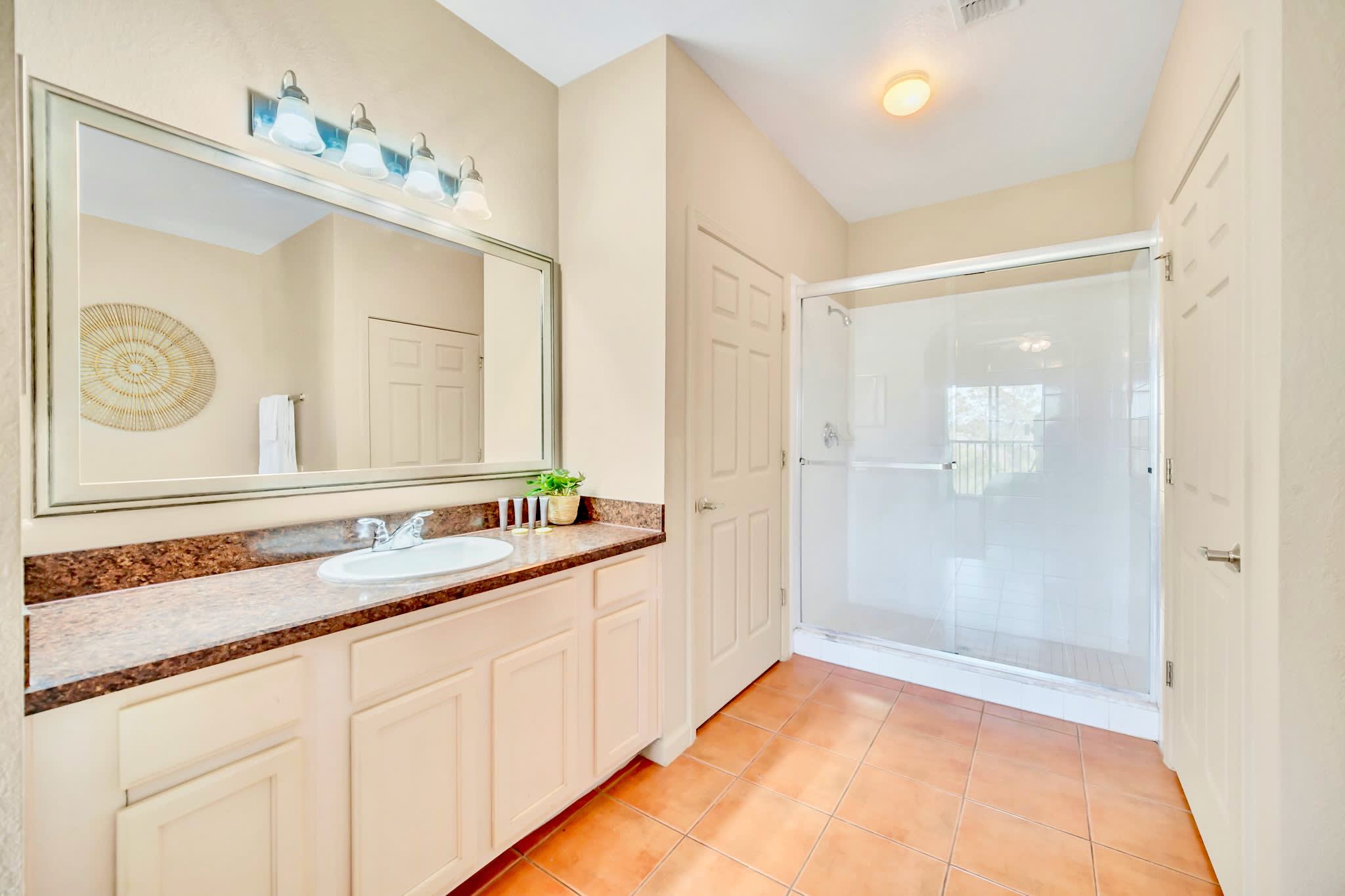 Bright bathroom with a single vanity featuring a light cabinet and granite countertop, a large mirror, and a glass-enclosed walk-in shower. The floor is tiled in a warm terracotta color.