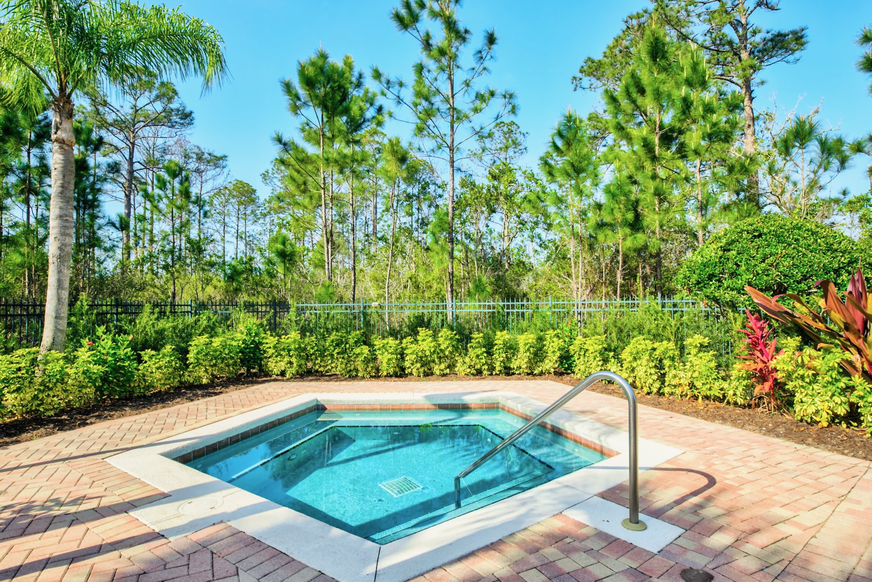 A square hot tub with sparkling blue water is set into a brick-paved deck, surrounded by vibrant green landscaping. Tall pine trees are visible in the background under a clear, sunny sky.