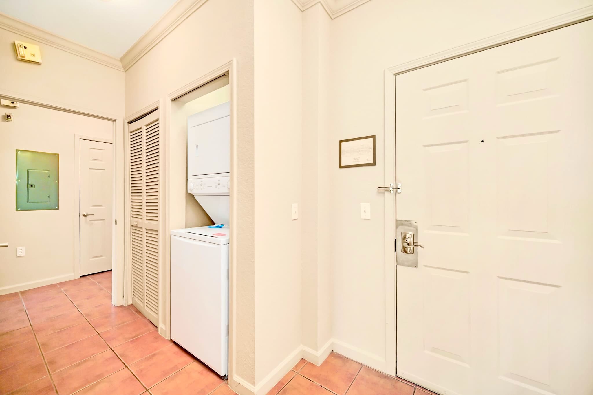 A functional entryway features a white front door and a stacked white washer and dryer unit neatly integrated into a closet. The space has light beige walls and terracotta-colored tile flooring.