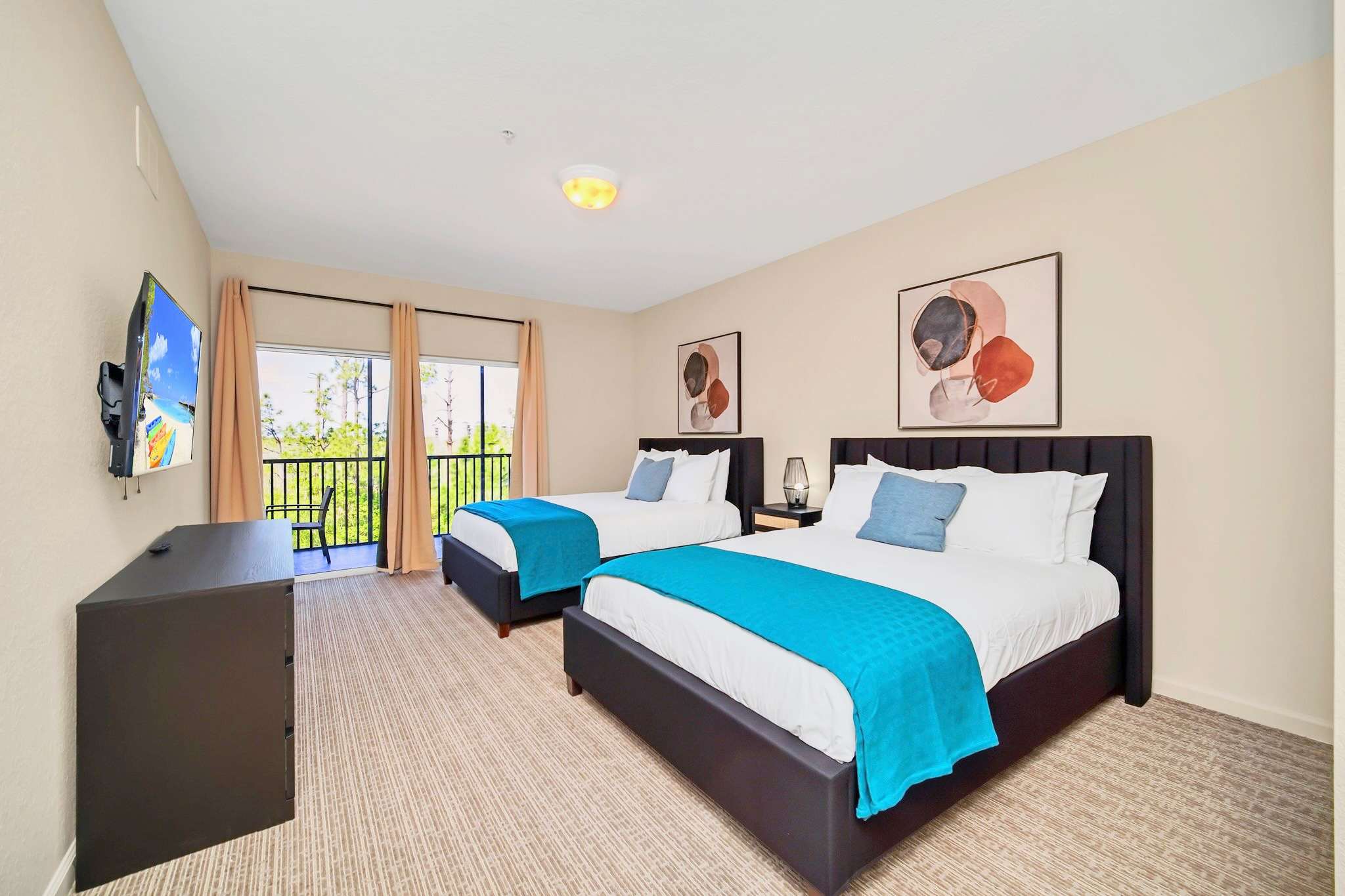 A bright bedroom features two beds with teal throws, light-colored walls, and a private balcony overlooking a natural landscape. The room is equipped with a wall-mounted TV and modern abstract artwork.