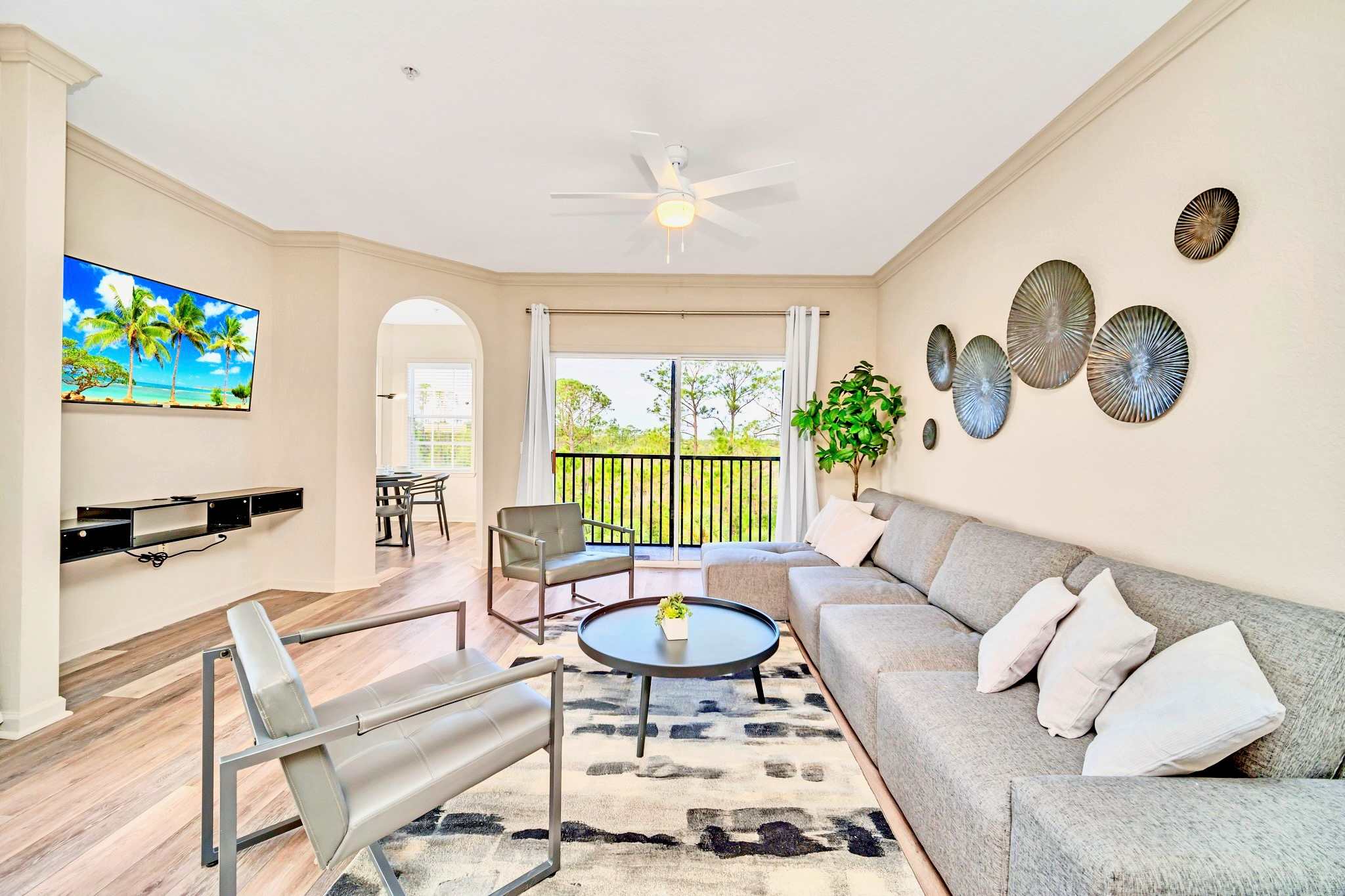 Spacious living room with a large gray sectional sofa, two modern gray armchairs, and a round black coffee table on a patterned rug. The wall features decorative metal plates and a potted plant, with sliding glass doors leading to a balcony.