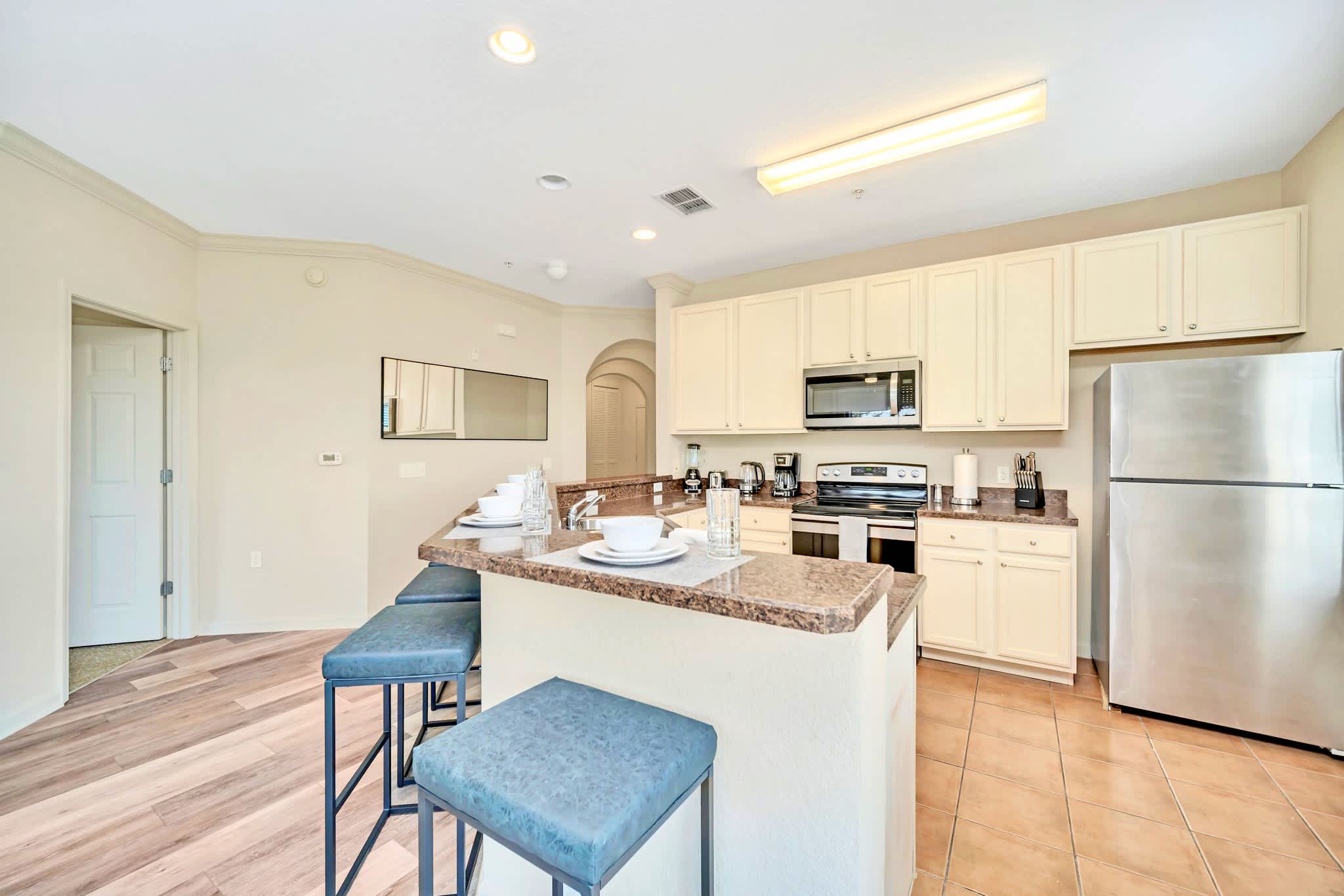 A bright kitchen features cream-colored cabinets, granite countertops, and stainless steel appliances, with a central island serving as a breakfast bar with three blue upholstered stools.