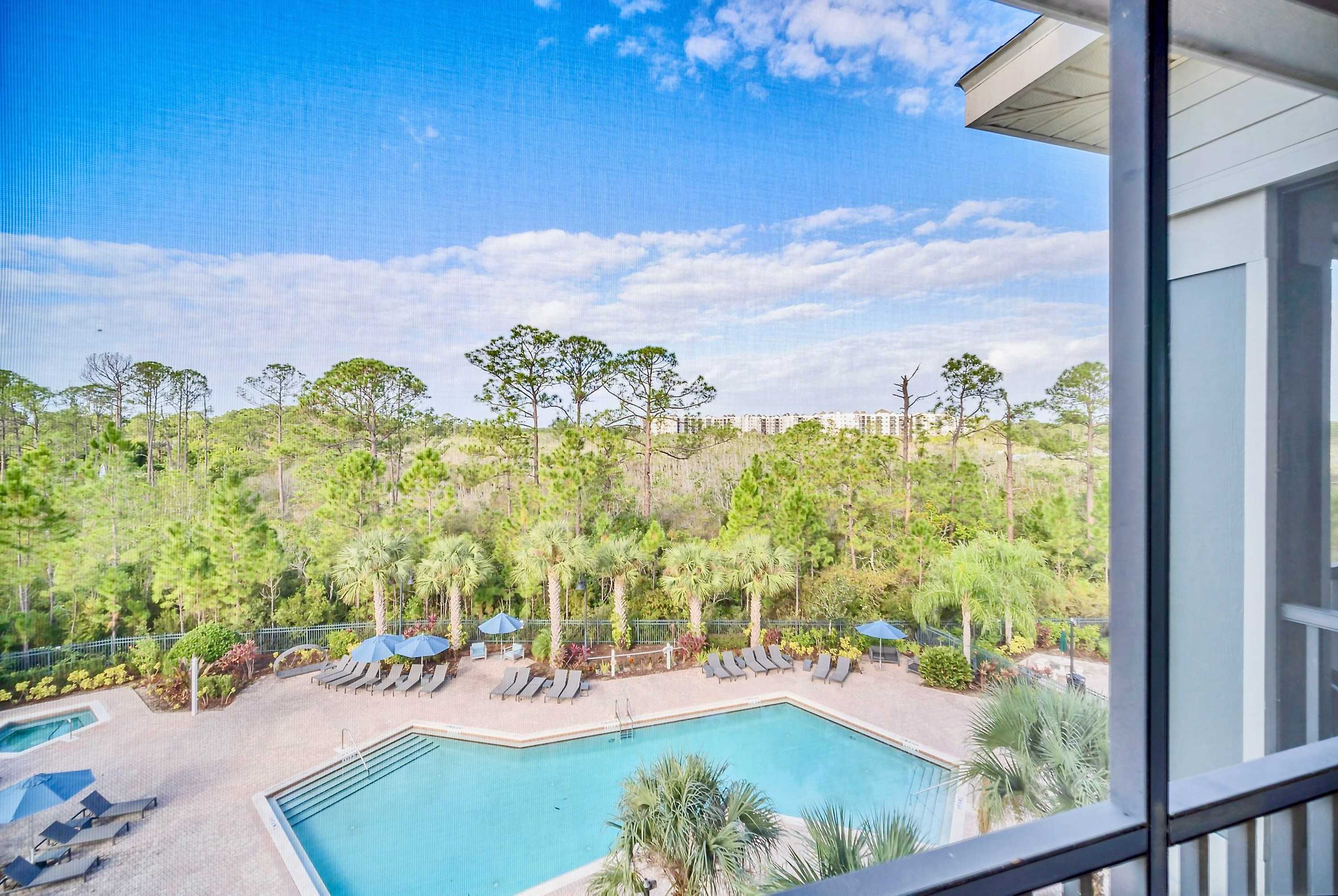 An elevated view of a communal outdoor swimming pool surrounded by lounge chairs, palm trees, and lush greenery, with a clear blue sky overhead.