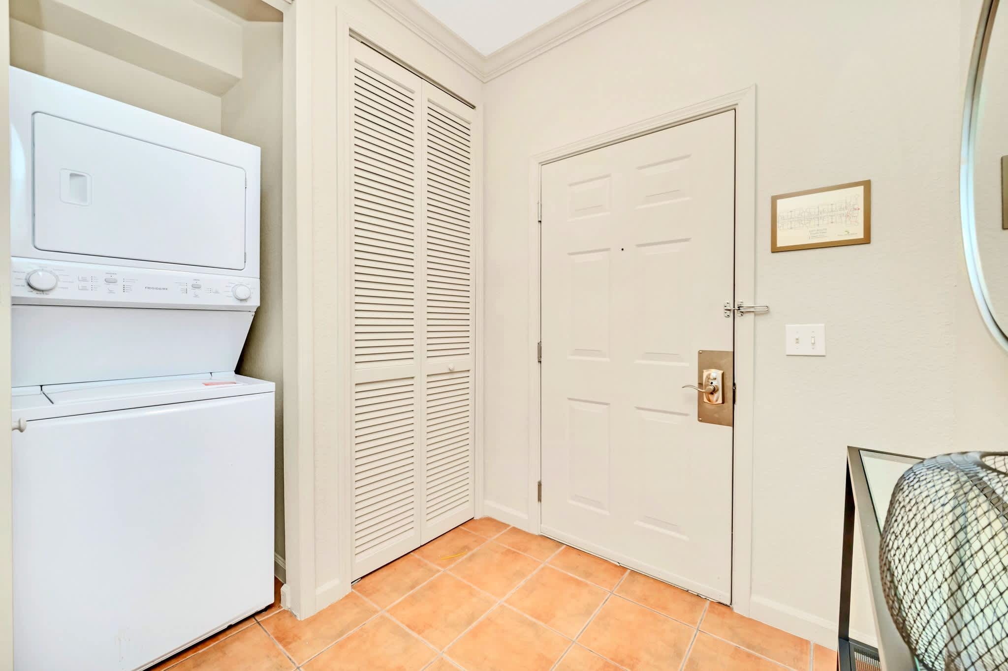 A compact laundry area features a stacked white washer and dryer unit next to a bi-fold closet door, with terracotta-colored tile flooring and a white entry door visible.