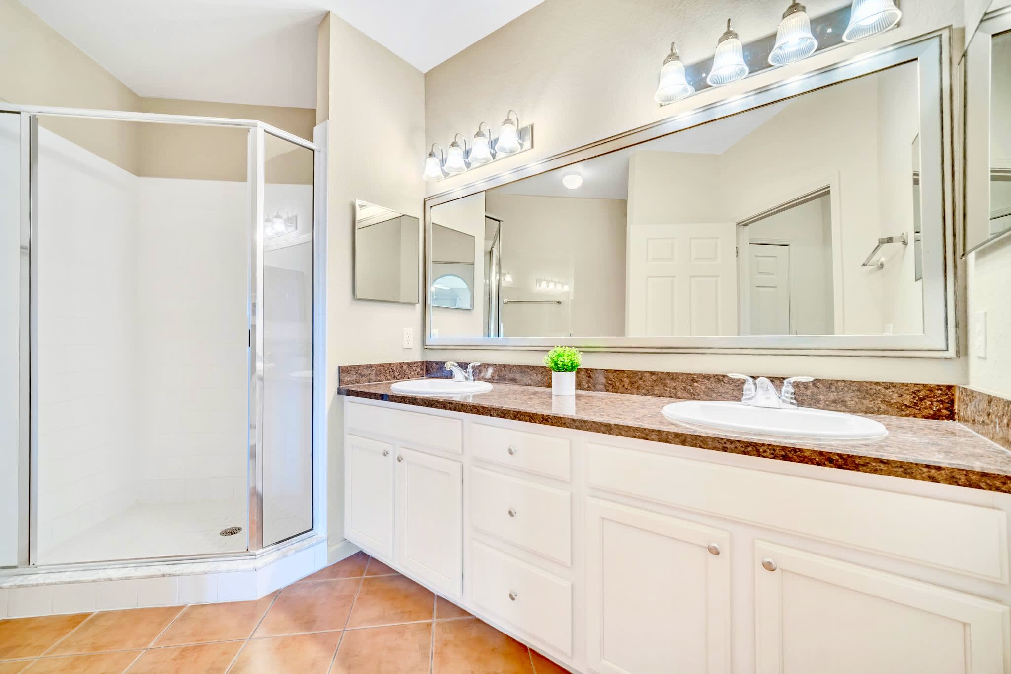 A spacious bathroom with terracotta-colored tile flooring features a long vanity with double sinks, a large mirror, cream cabinets, and a walk-in glass shower enclosure.
