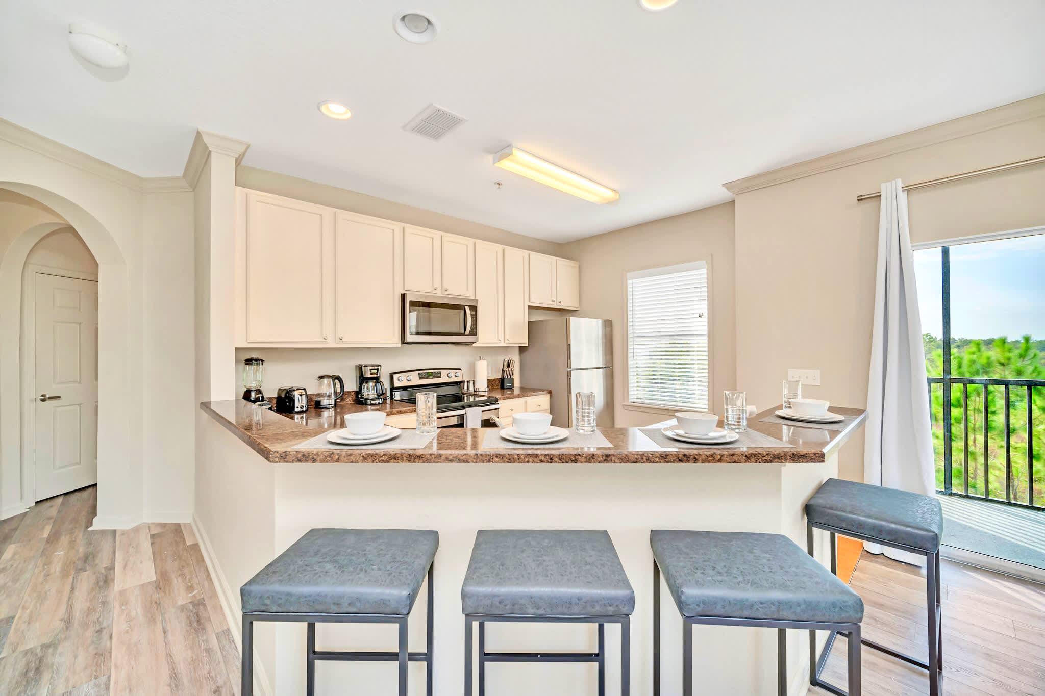 View of the modern kitchen from the living area, showcasing a breakfast bar with three stools and a double sink. Light-colored cabinets and stainless steel appliances are visible, with a window providing natural light and a glimpse of the outdoors.