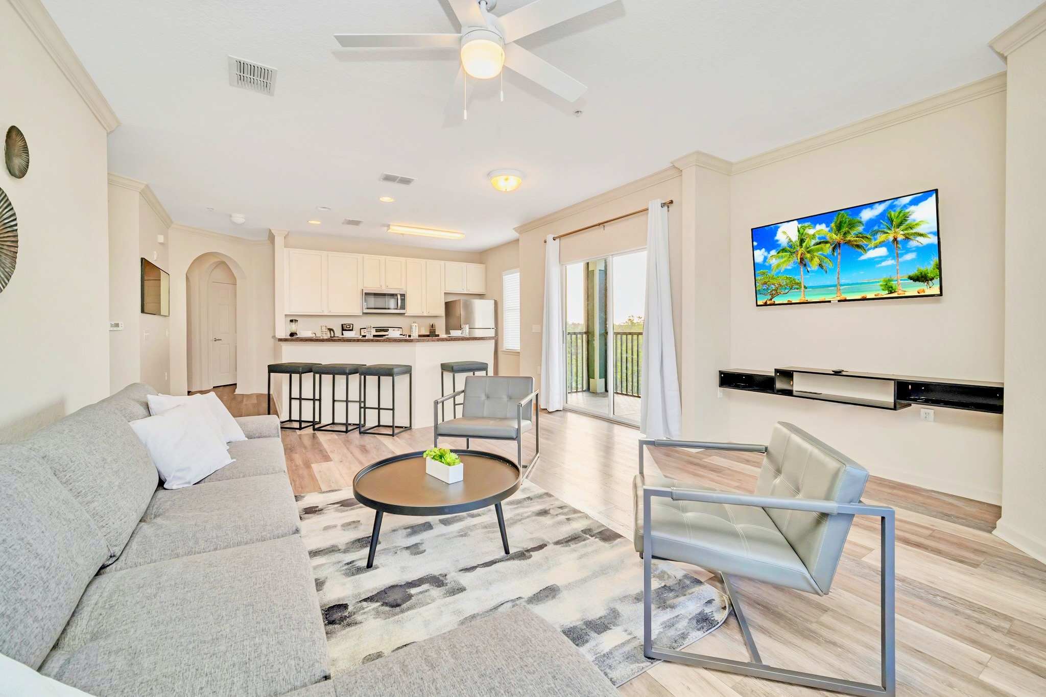 Open-concept living room featuring a gray sectional sofa and armchairs, with a wall-mounted TV. The adjacent kitchen's breakfast bar with stools is visible, creating a seamless flow between the spaces and offering balcony views.
