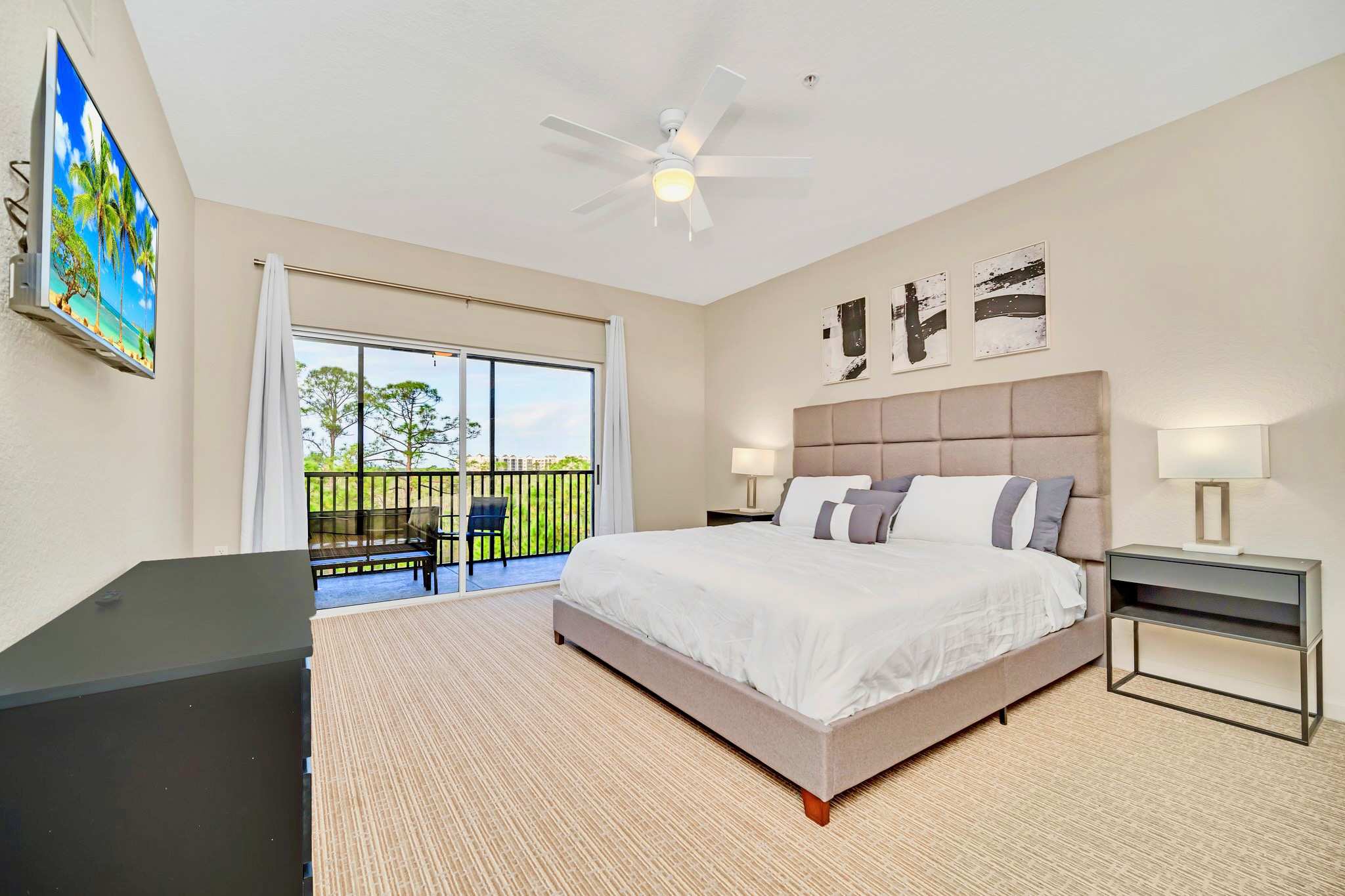 A spacious bedroom with light beige walls and carpet features a large gray upholstered king-size bed with white and gray bedding, two dark nightstands with lamps, and a flat-screen TV mounted on the wall. A sliding glass door leads to a balcony.