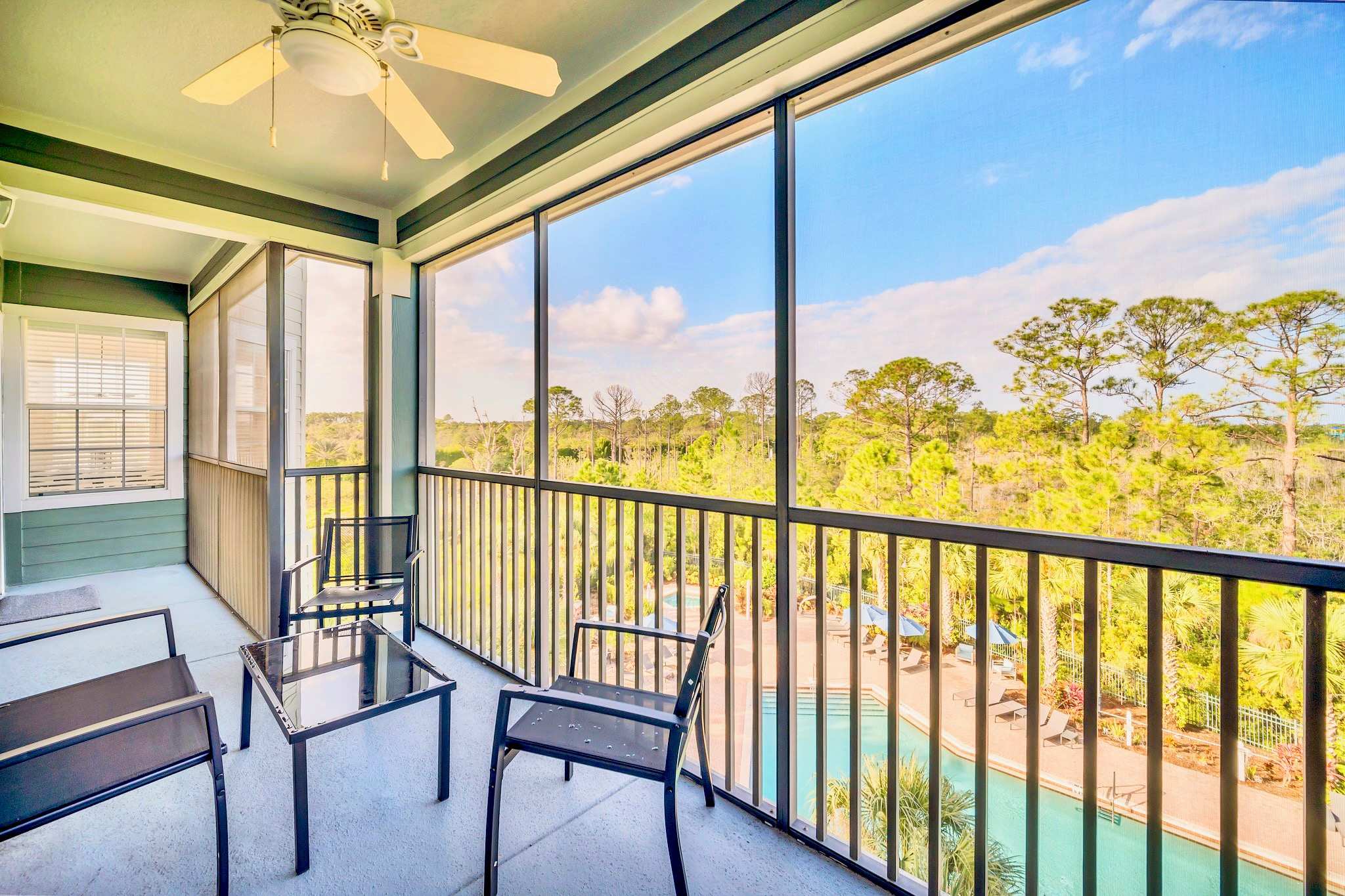 A screened-in balcony with a ceiling fan offers outdoor seating with a glass-top table and chairs, overlooking a lush green landscape and a swimming pool below.