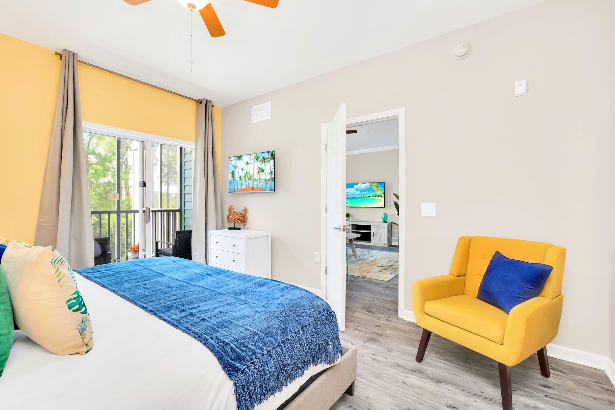 A bright bedroom with a yellow accent wall, a large bed with a blue throw, a yellow accent chair, and a wall-mounted TV, featuring light grey wood-look flooring and access to a balcony through sliding glass doors.