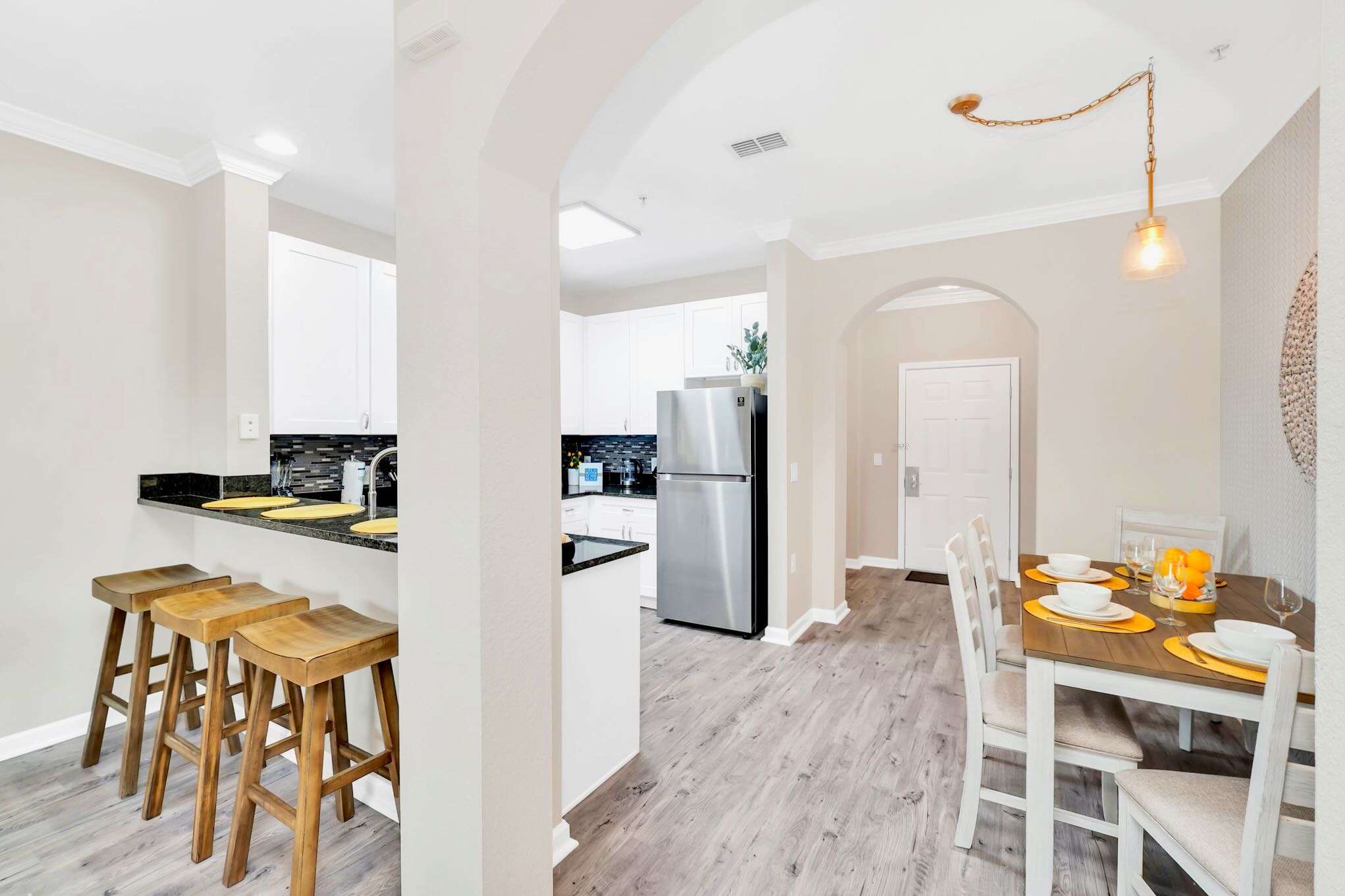 A bright open-concept space featuring a dining area with a wooden table and white chairs, leading into a modern kitchen with white cabinets, dark countertops, and stainless steel appliances, all set on light grey wood-look flooring. An arched doorway leads to the front entrance.
