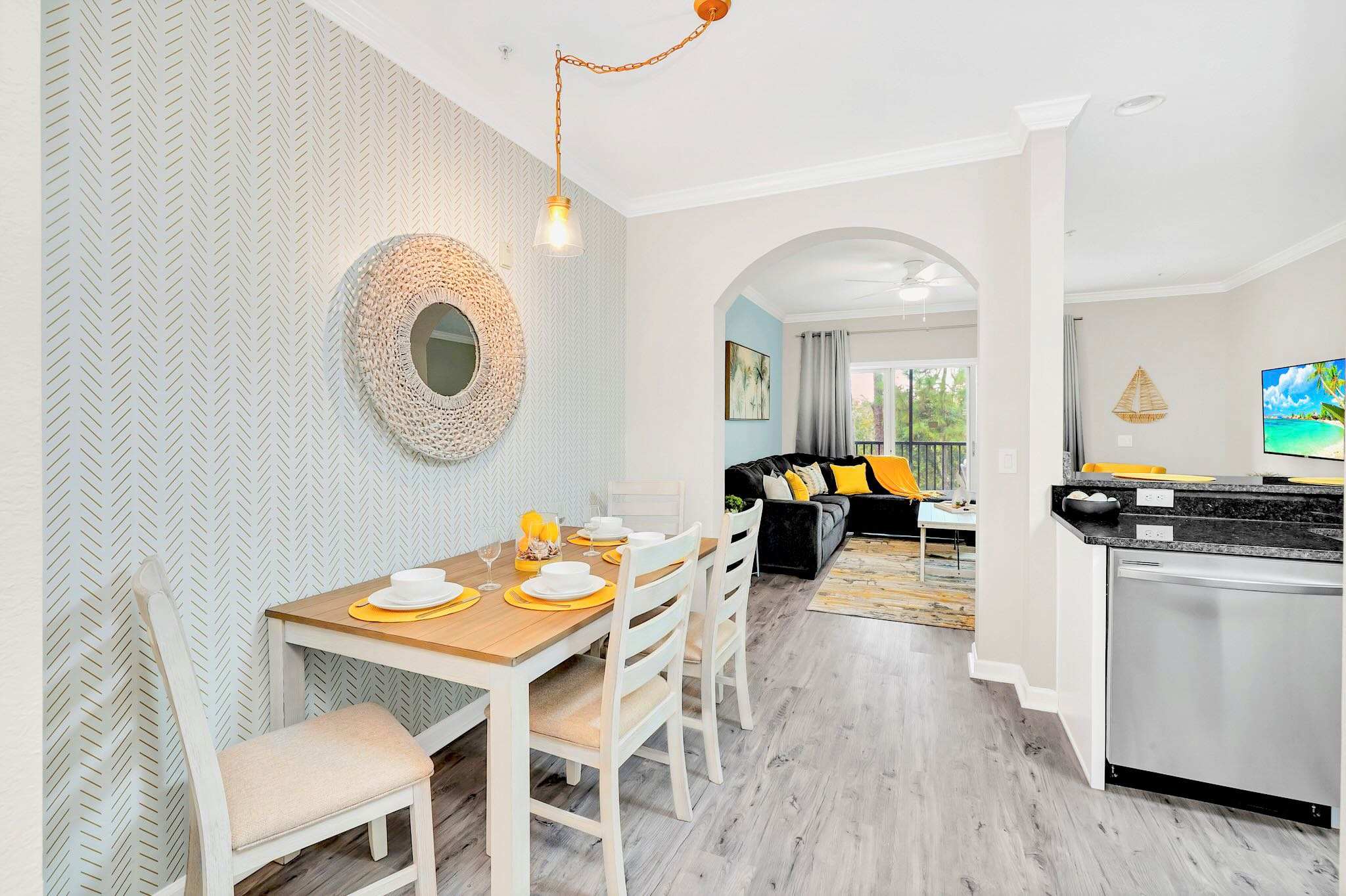 Dining area with a light wood table and four chairs, featuring a patterned accent wall with a woven mirror, and an open view towards the kitchen and living room.
