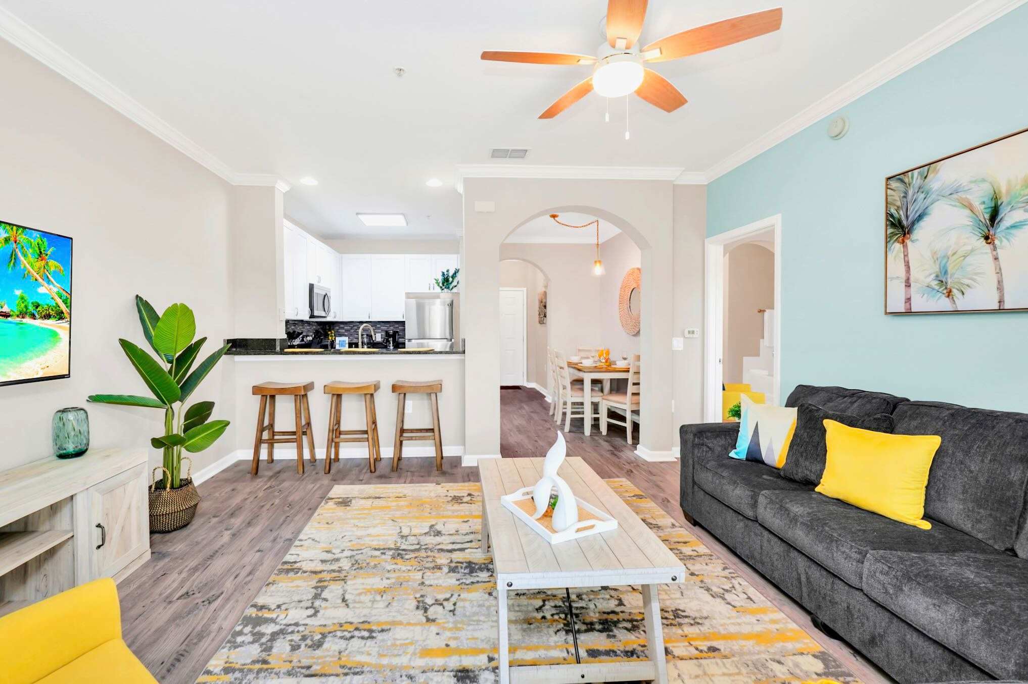 Open-concept living space showcasing a gray sectional sofa, a kitchen island with three wooden bar stools, and a dining area beyond an arched doorway, all connected by light wood flooring.