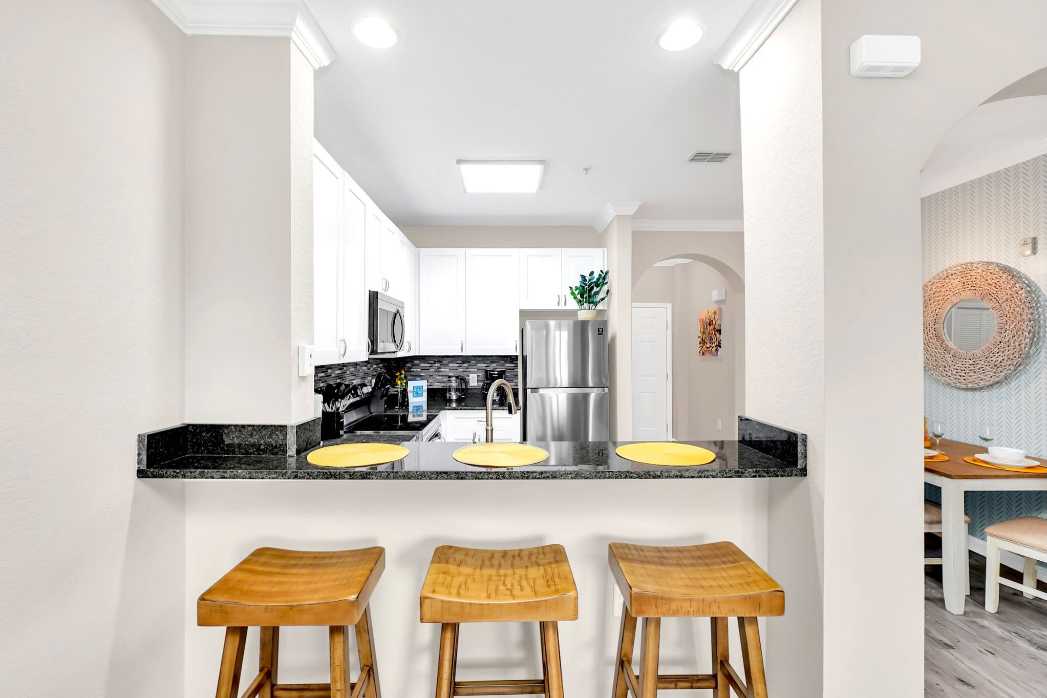 A kitchen bar area with dark granite countertops and three wooden bar stools, separating the kitchen from the dining space, featuring light grey wood-look flooring and a view towards an arched entryway.