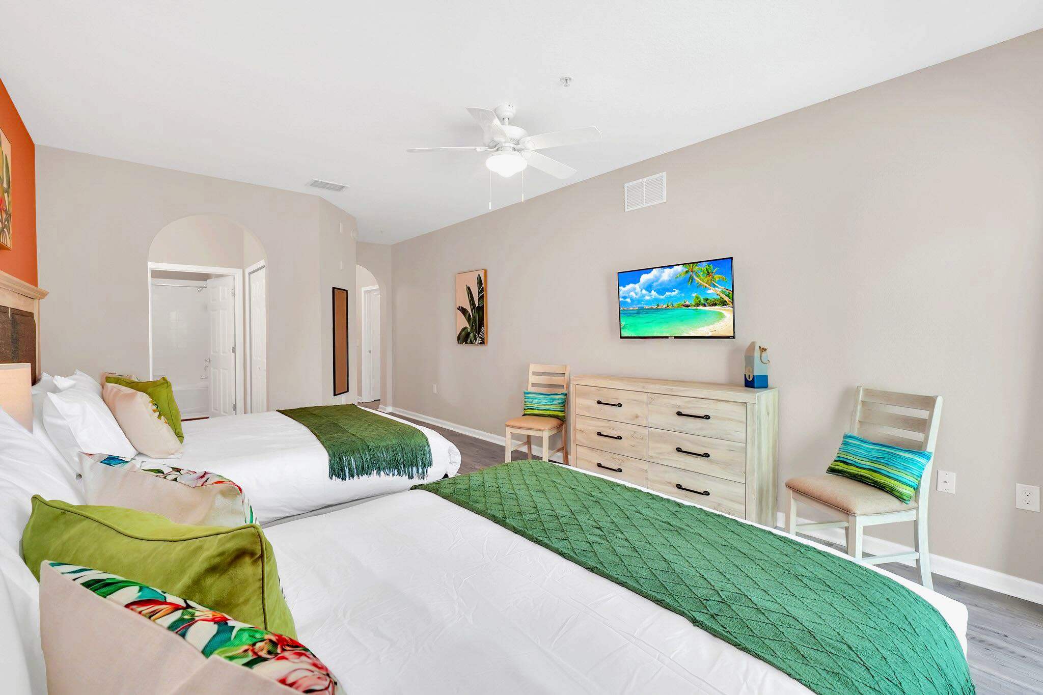 A spacious bedroom featuring two queen beds with green throws, light grey wood-look flooring, and a dresser with a wall-mounted TV, with an arched doorway leading to an ensuite bathroom.