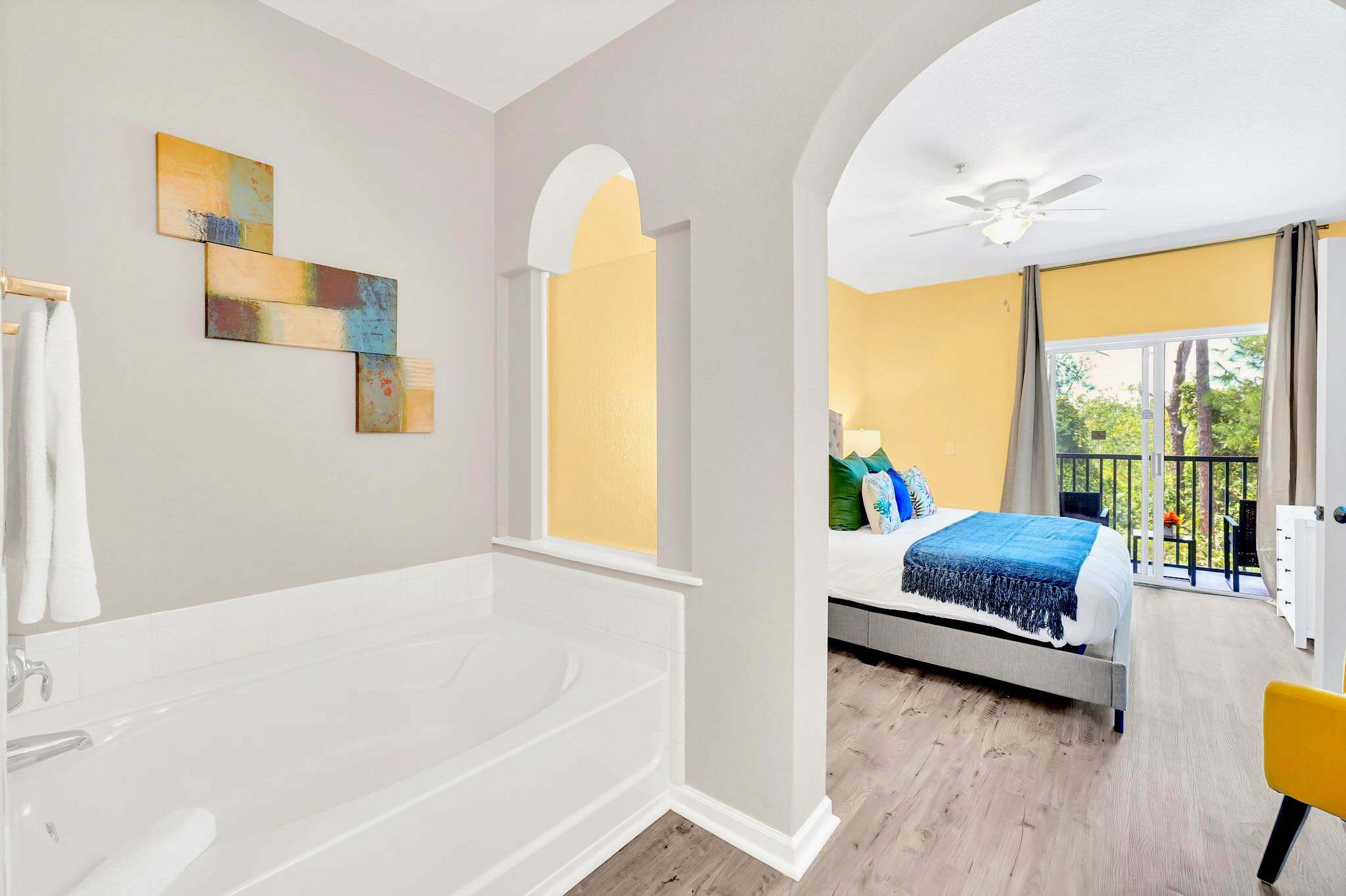 A master bedroom with a yellow accent wall and a bed with blue bedding, featuring light grey wood-look flooring and an open archway leading to an ensuite bathroom with a built-in bathtub.